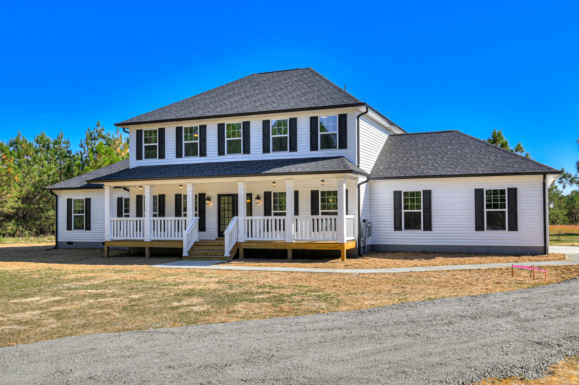 White siding house with black shingle roof, black shuttered windows, white framed windows, wooden deck with white railing, red tape on fence, surrounded by trees under blue sky.