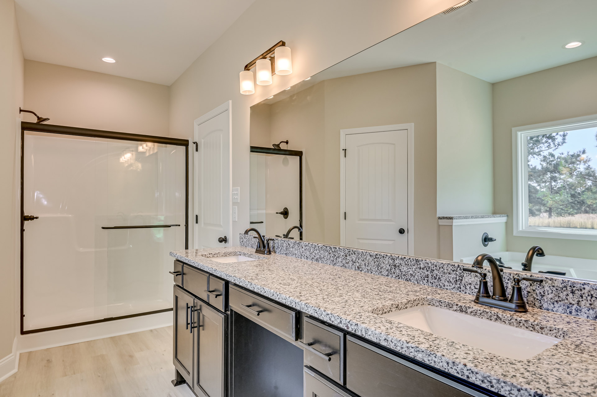 Bathroom with double vanity featuring white cabinets and chrome faucets, glass-enclosed shower, large mirror, tiled walls, window overlooking trees, and modern light fixtures with