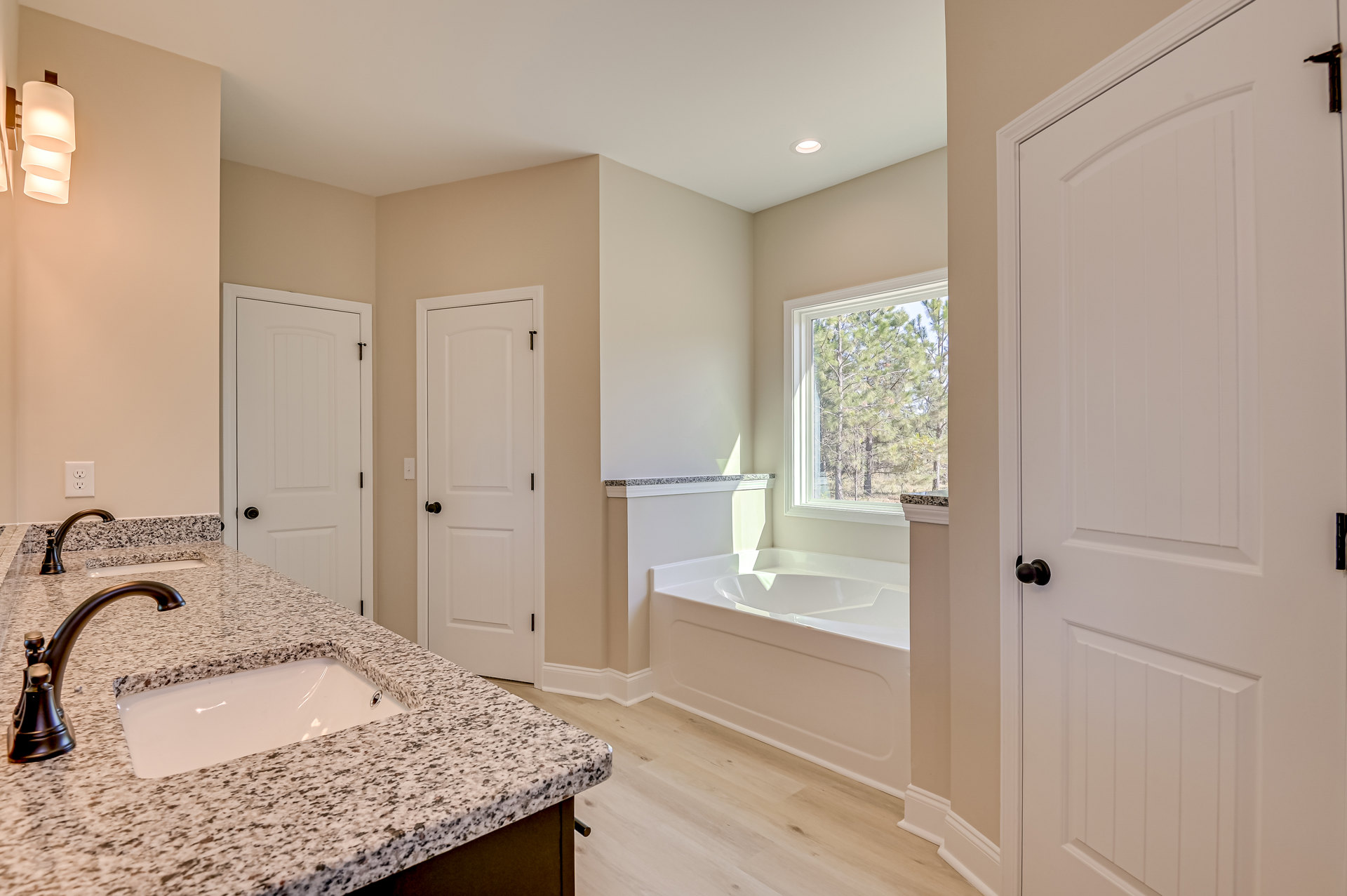 Bathroom featuring a marble countertop with undermount sink and chrome faucet, freestanding bathtub near a window overlooking trees, white door with black knob, and modern lamp