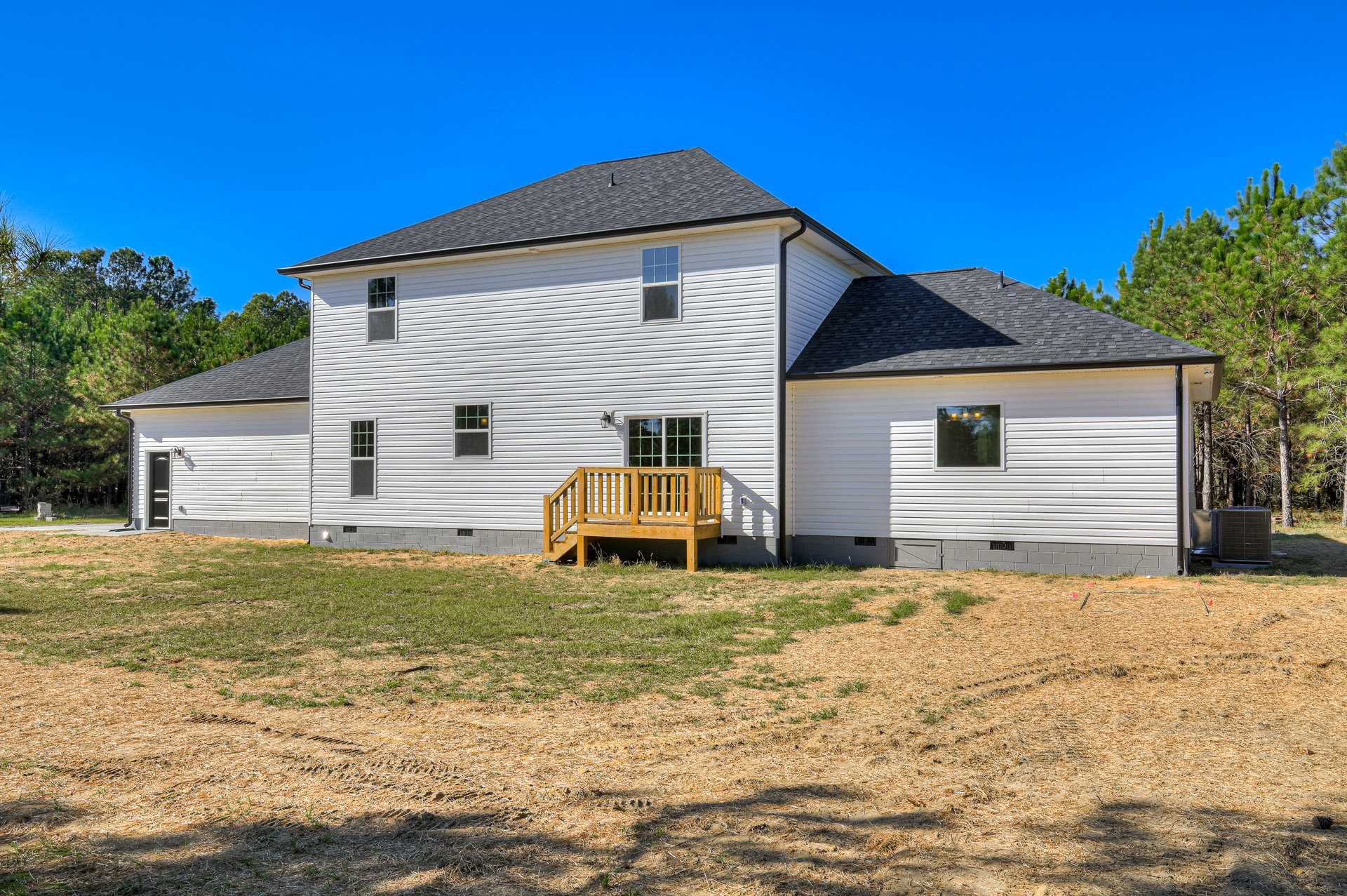 Two-story house with white siding, multi-pane windows, wooden porch and deck, stairs leading to a green lawn, surrounded by trees and landscaping.