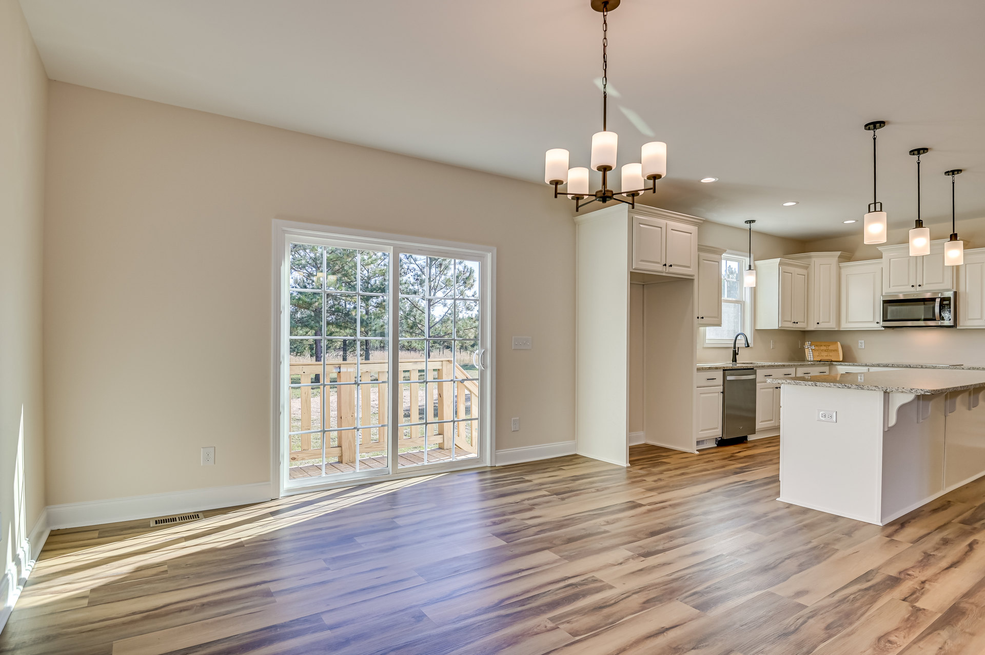 Open kitchen and living room with wood flooring, marble-topped island, white cabinetry, stainless steel microwave and dishwasher, large window overlooking deck and trees, patio