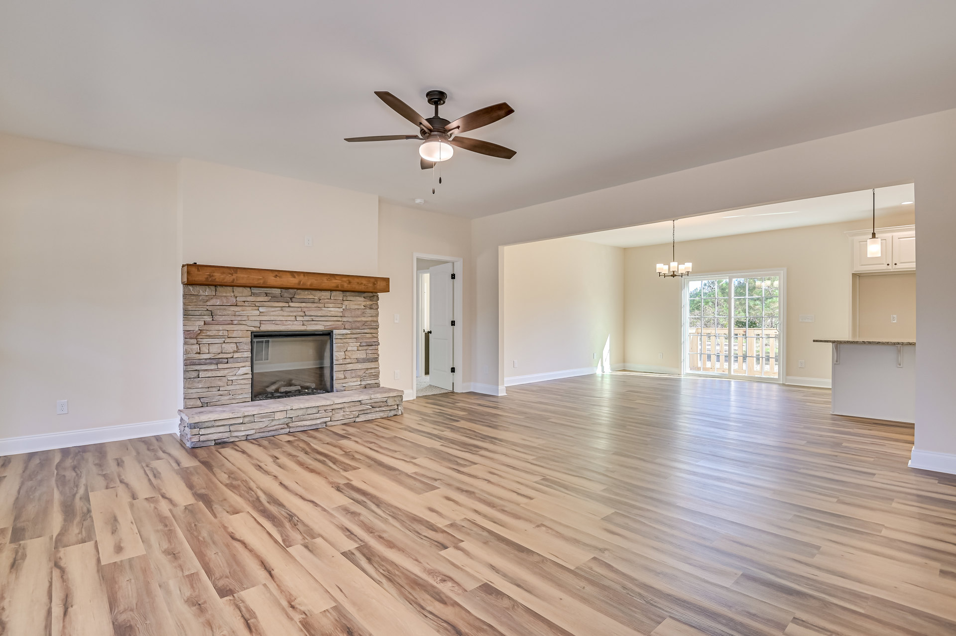 Living room with hardwood floor, ceiling fan with light, fireplace filled with wood logs, window overlooking trees, and white door with black handle