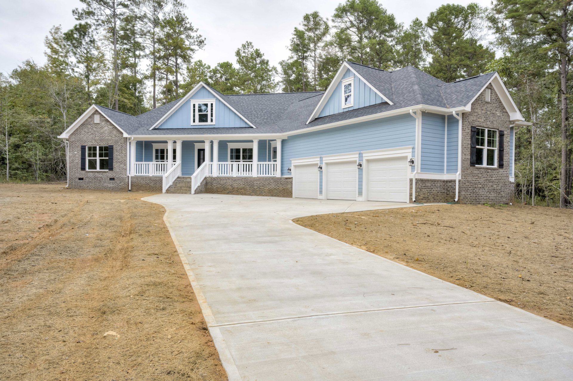 Blue house with white trim, large front window with white frame, concrete driveway bordered by green lawn, small porch, mature trees in background, clear sky overhead