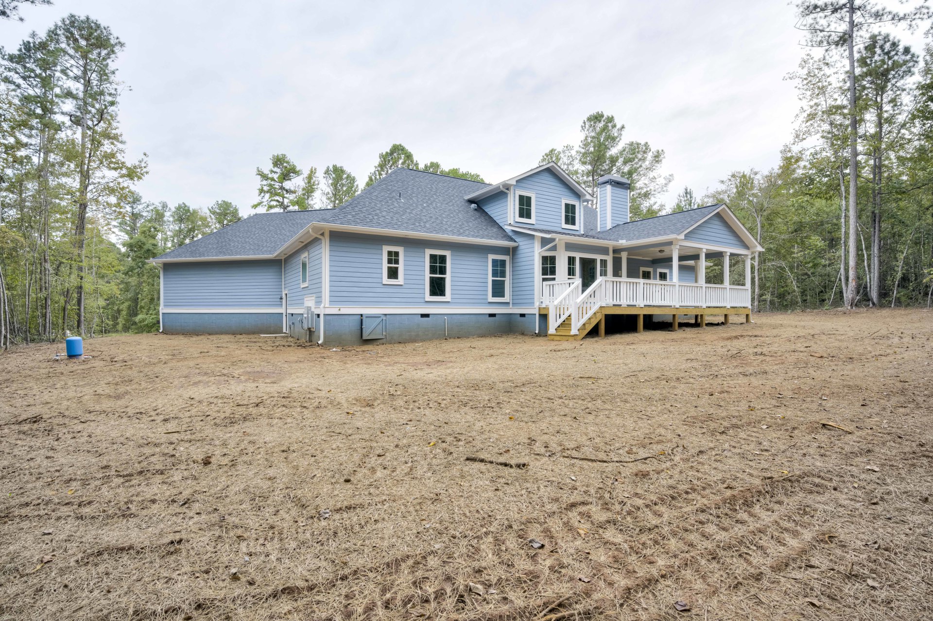 White porch with staircase leading to entry, expansive green yard, paved driveway, mature trees bordering property, light-colored siding, cloudy sky overhead