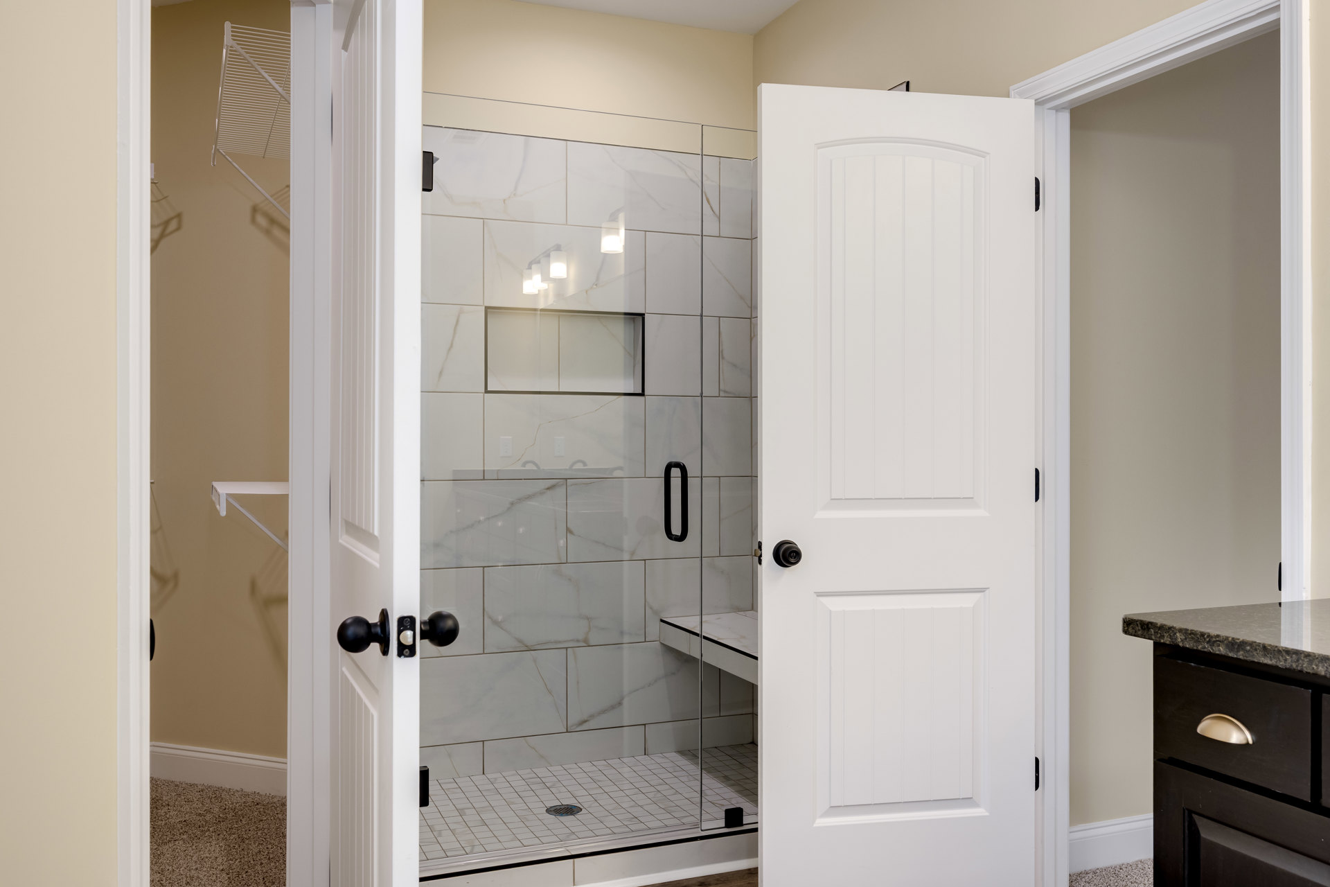 Walk-in shower with clear glass door, gray tile floor with linear drain, adjacent white door featuring black knob, vanity drawer with gold handle, wall-mounted mirror, black