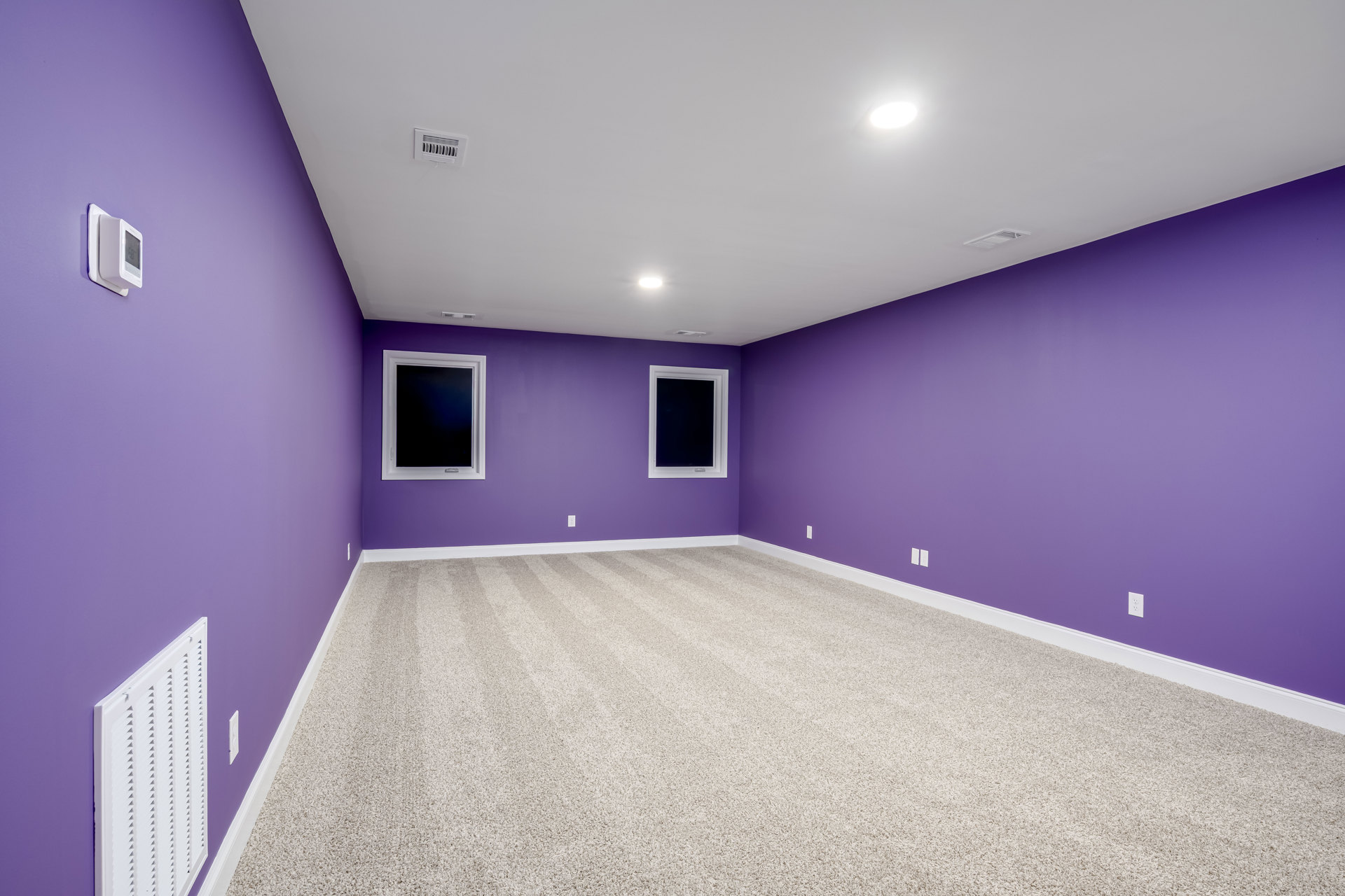 Purple-walled room with matching carpet, white-framed window with black screen, white ceiling vent, bright ceiling light, and white electronic thermostat on the wall