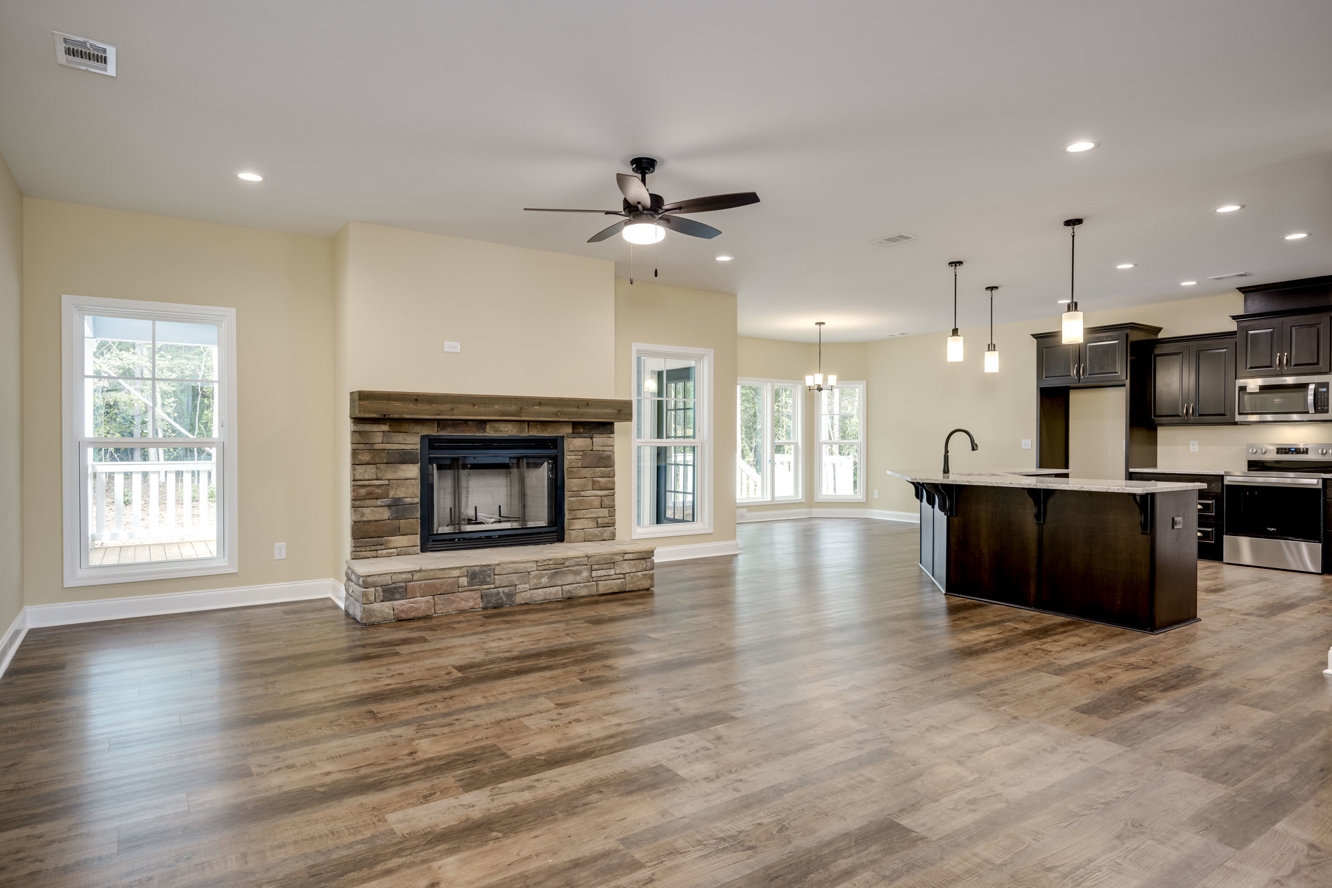 Spacious open living area featuring wood flooring, central fireplace with mesh screen, ceiling fan with light, large windows framed by white railing overlooking trees