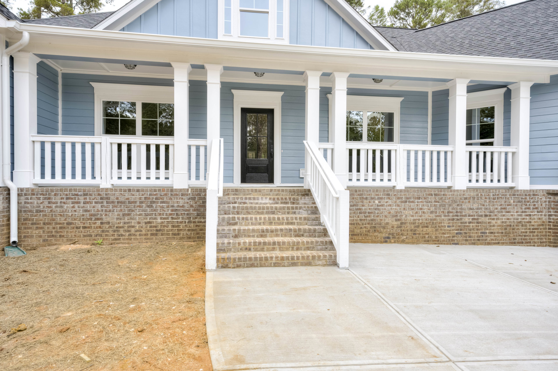Front porch with white pillars, black door featuring glass window, concrete stairs and patio, white railing, brick wall accents, gray siding, tree visible behind roof