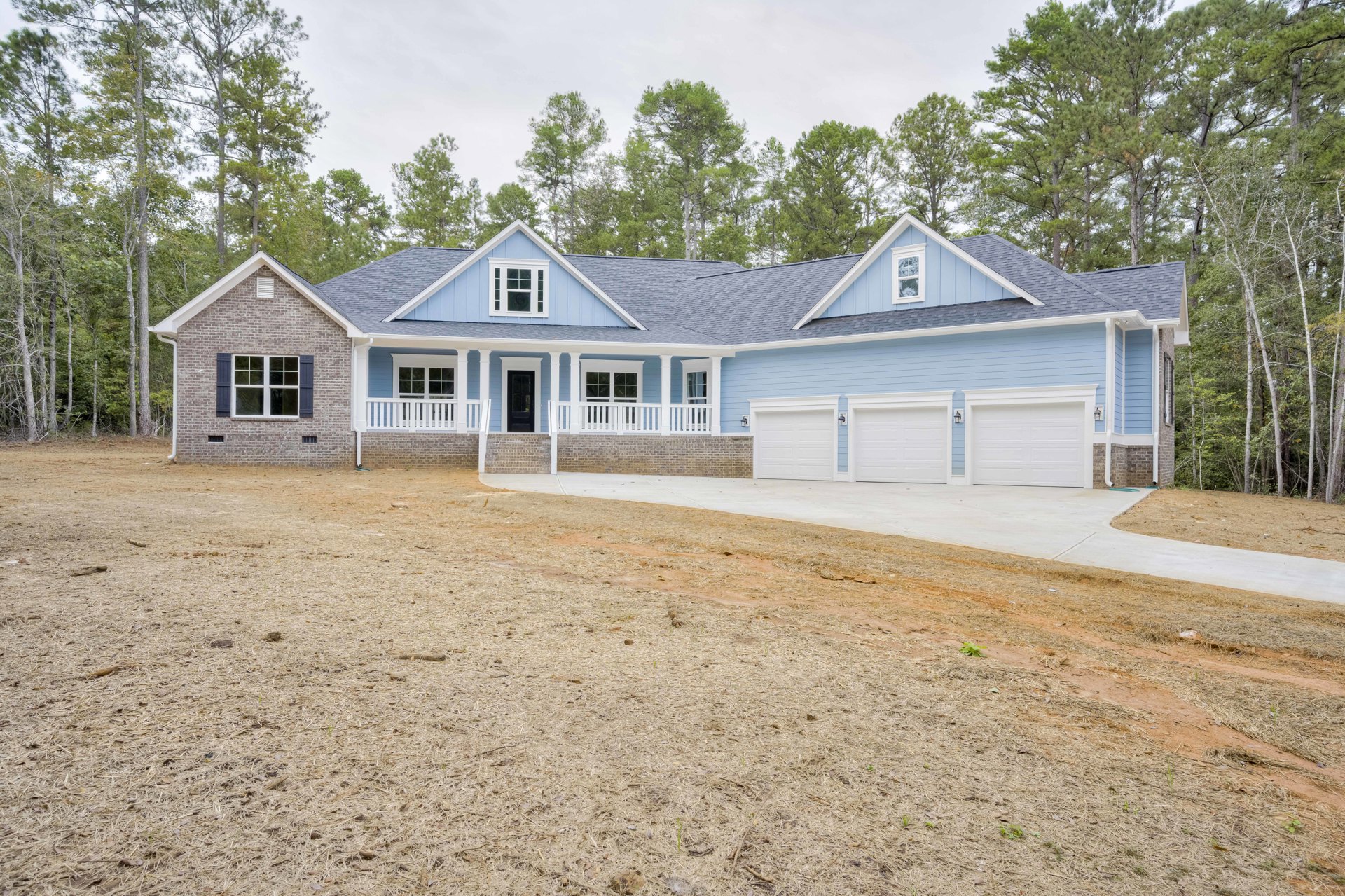 Blue house with white trim and pillars, white railing on porch, brick driveway bordered by brick wall, mature trees surrounding property, large windows with white frames