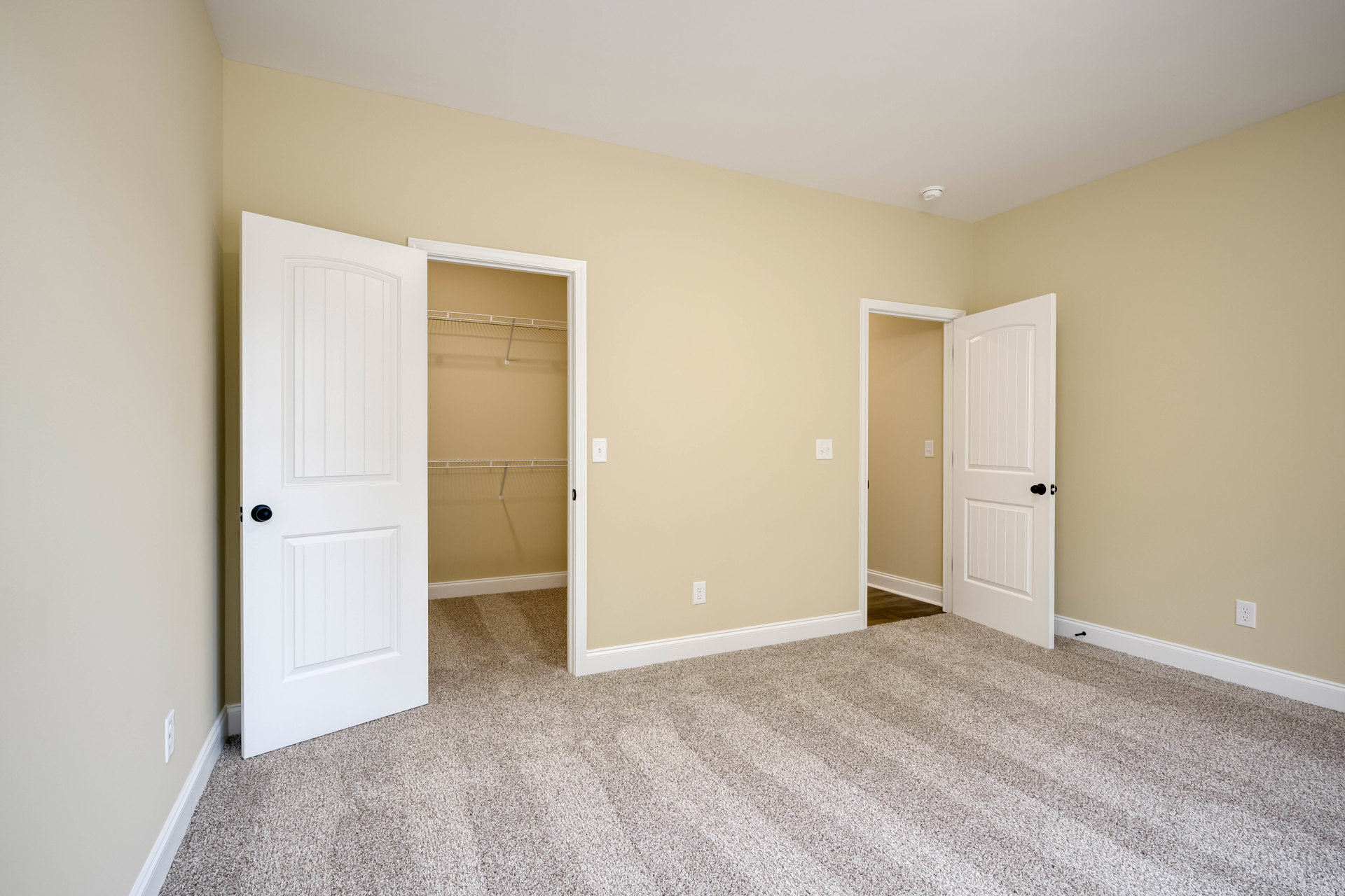 Bedroom with beige carpet, two white doors open, black door knobs, light switch on wall, closet with white shelf and open door, white baseboards and molding