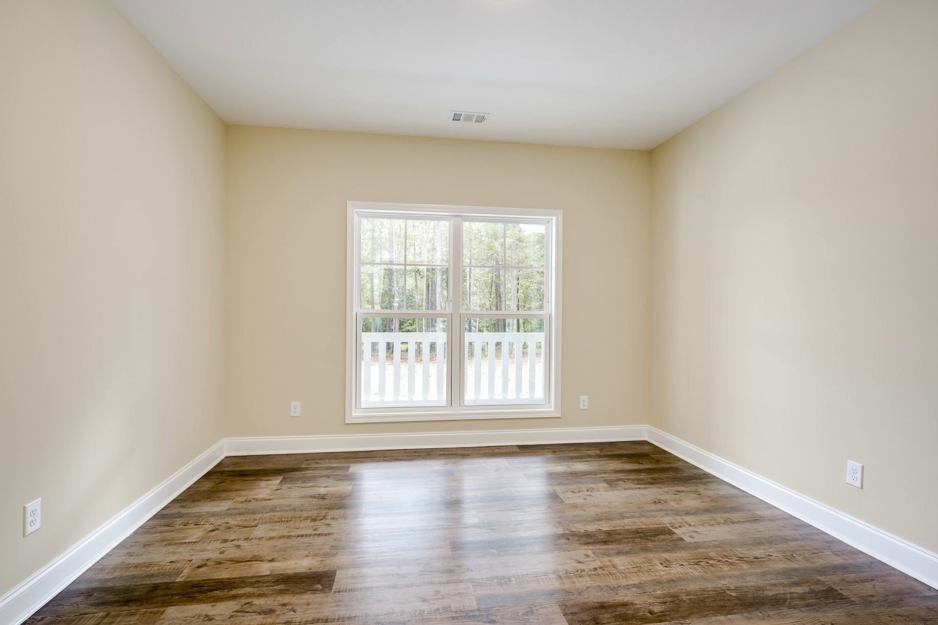 Sunlit room featuring a double white-trimmed window with a white railing, hardwood flooring, white baseboards, and a ceiling vent; green trees visible outside.