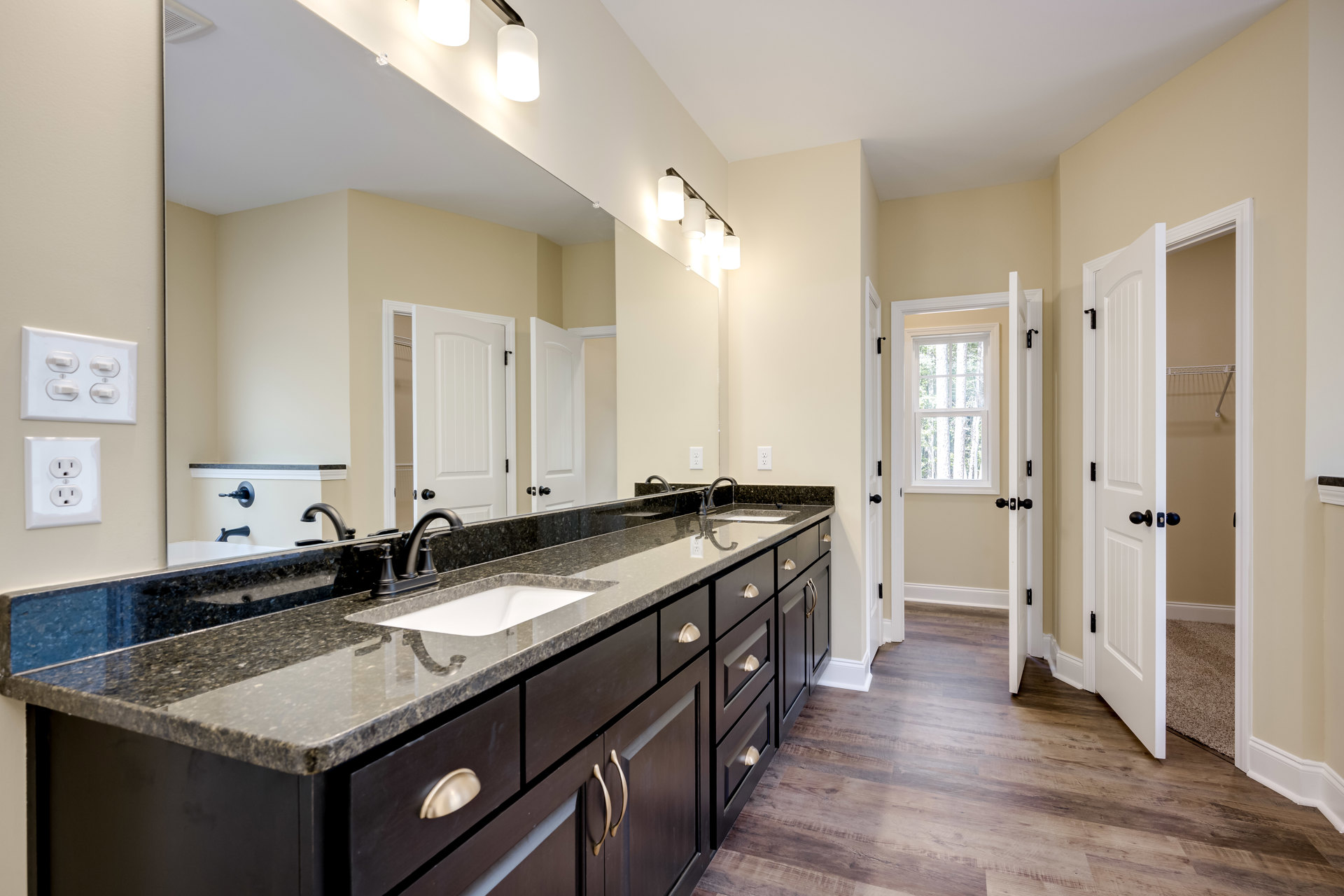 Bathroom with double vanity featuring white sinks, stone countertop, large framed mirror, wood cabinetry, tile flooring, and a window overlooking trees.