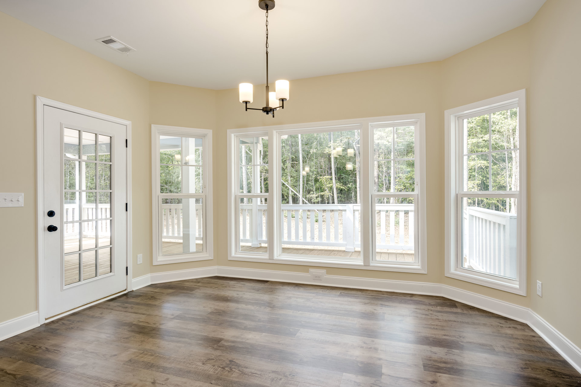 Hardwood floored living space featuring a glass-paneled door, multiple large windows with views of porch and trees, decorative molding, and a central chandelier.