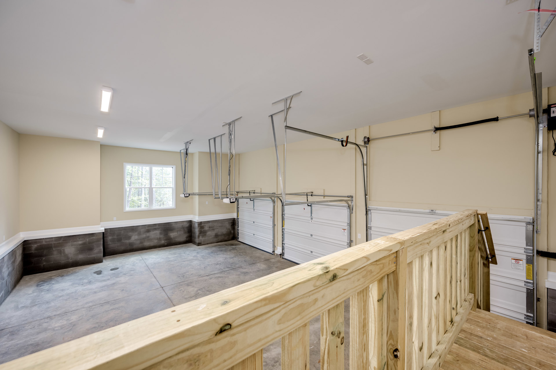 Garage interior featuring plaster walls, composite ceiling, concrete floor, and a wooden railing beside a metal-barred garage door; window reveals trees outside.