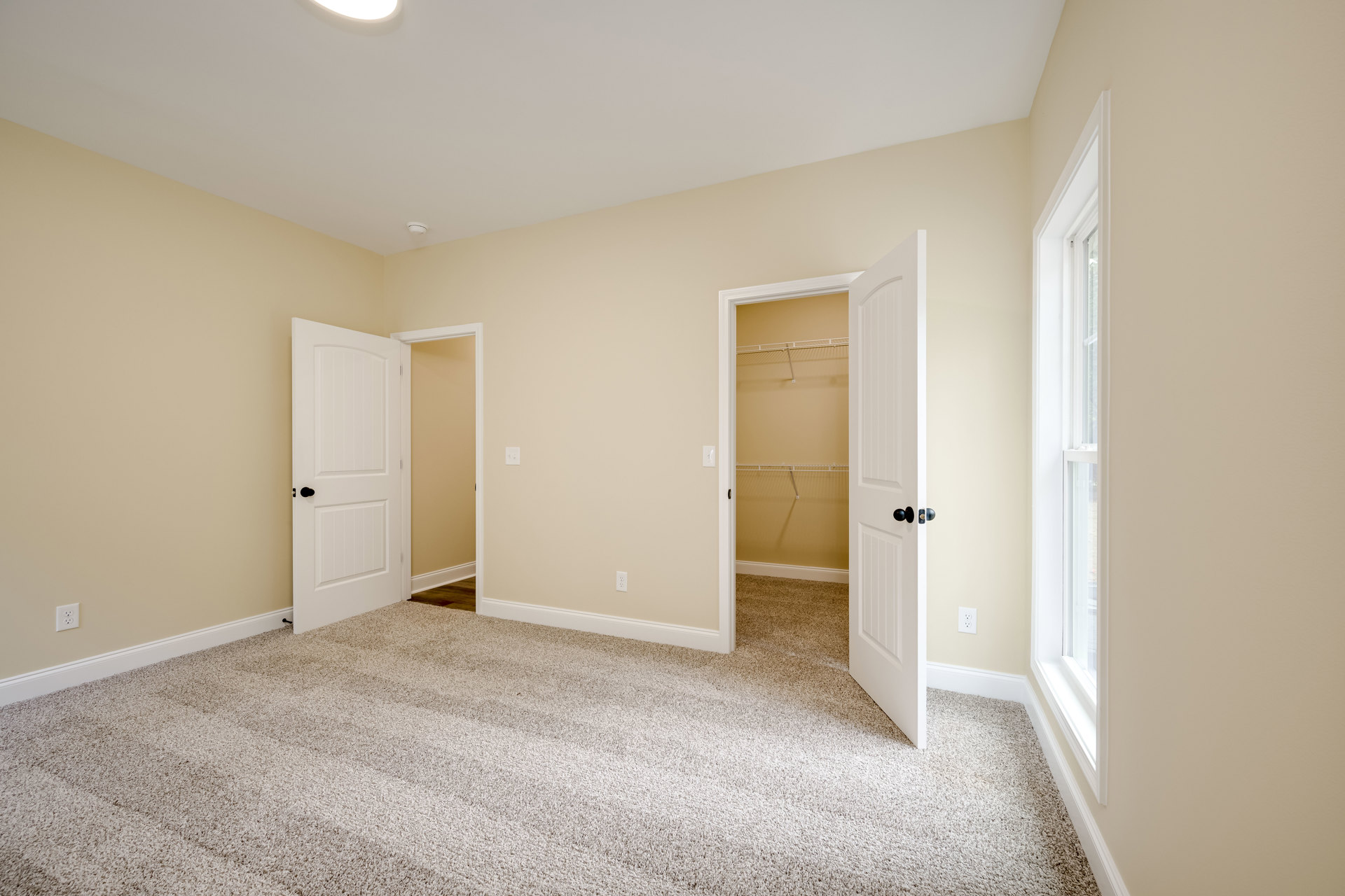 Tan-walled room with two open white doors featuring black handles, carpeted floor, white door frames, and a closet visible.