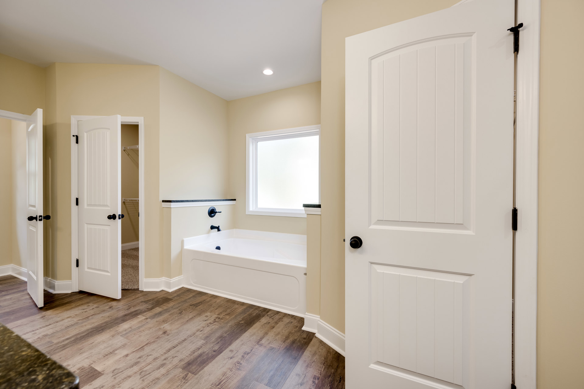 Bathroom with white freestanding bathtub and black faucet, wood plank flooring, white walls, marble countertop beneath window, white door with black knob