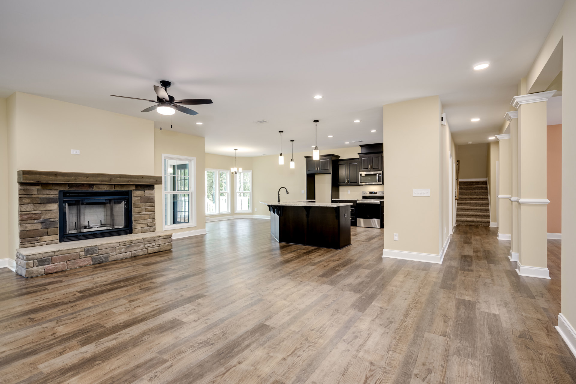 Open concept living room with wood flooring, central fireplace with screen, ceiling fan with light, and adjacent kitchen featuring black countertops