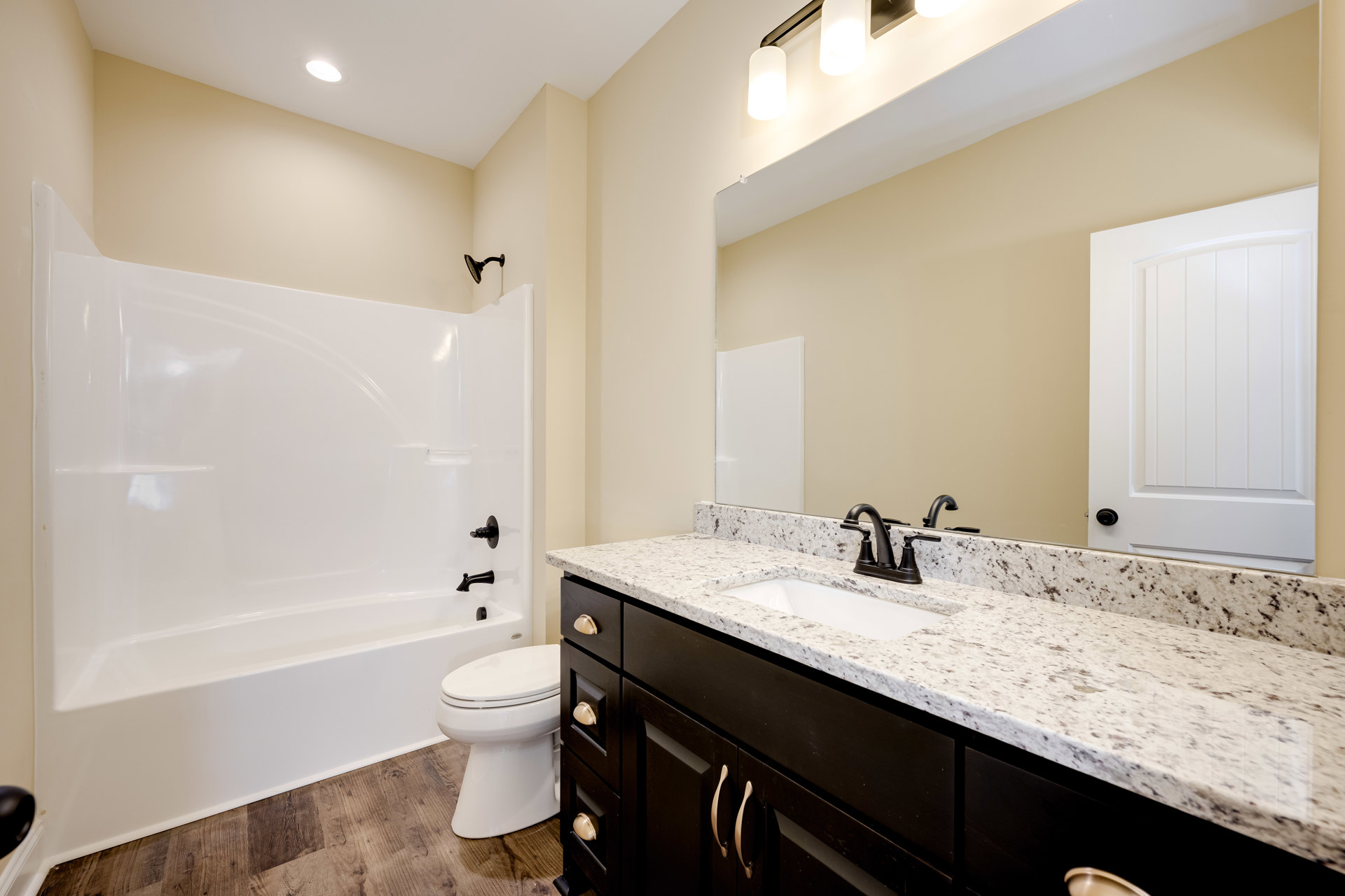 Bathroom with white bathtub, closed-lid toilet, rectangular sink on stone countertop, black-handled shower head, white door with curved top, light tile walls and floor, built-in