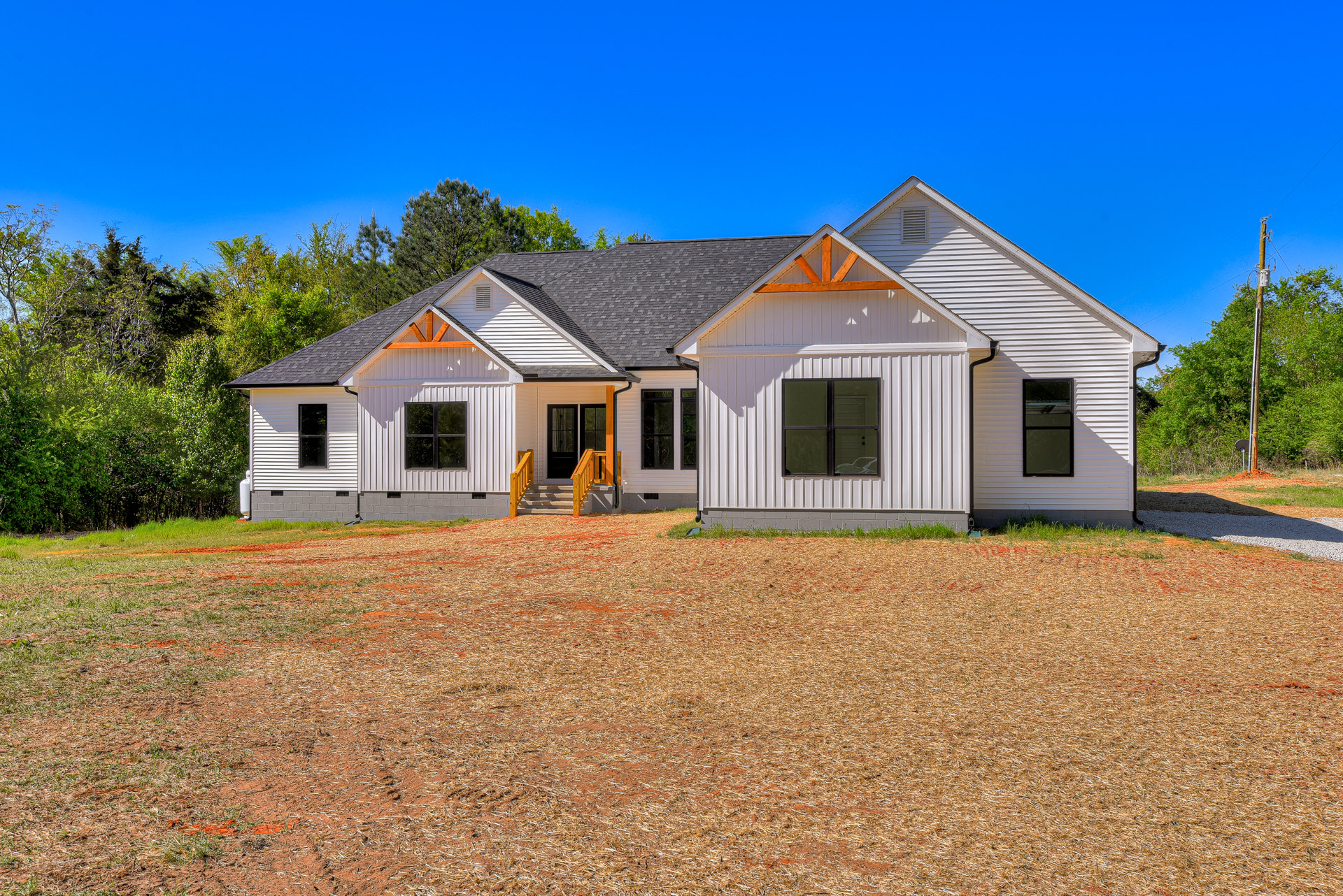Two-story house with black roof, black-framed windows, yellow stair railings, wood chip landscaping, paved driveway, and Robert Frost Farm visible in the background.