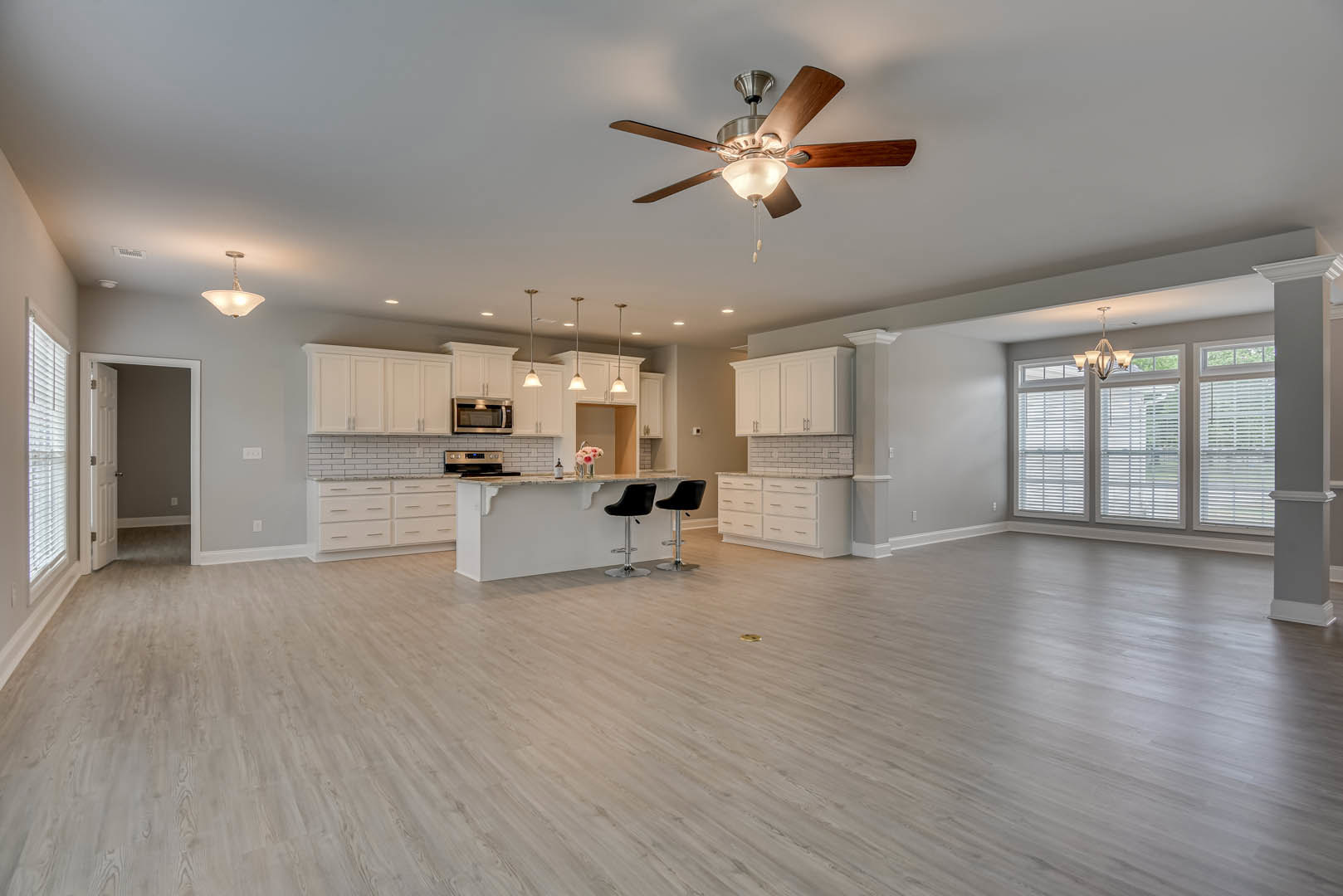 Spacious white kitchen featuring a ceiling fan with light fixture, white cabinetry, wood and laminate flooring, black bar stools with silver bases, and built-in microwave