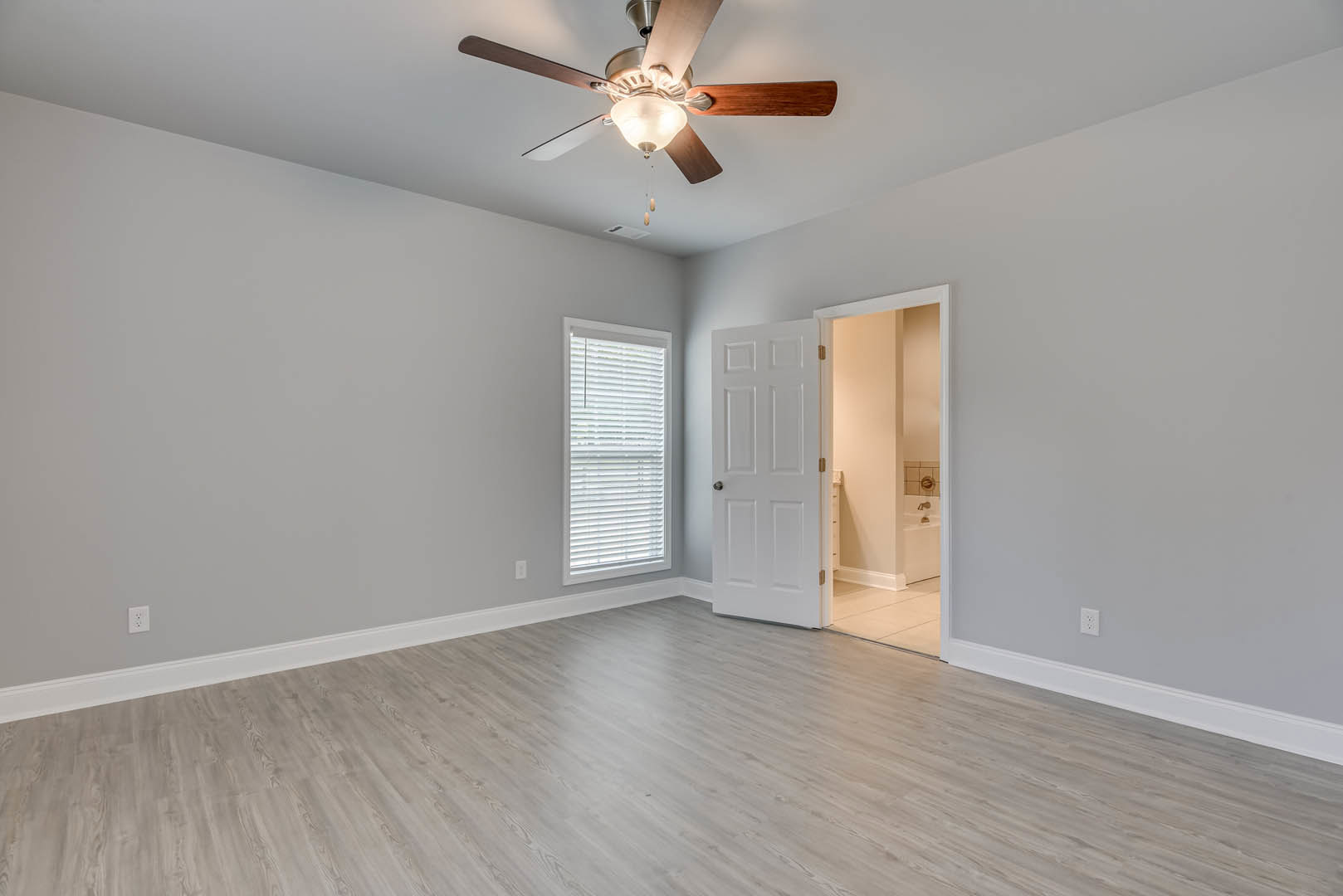 Ceiling fan with light fixture, hardwood flooring, white walls, window with blinds, white door with silver knob, partial view of bathroom with bathtub and sink