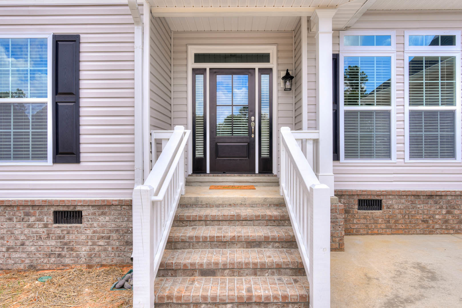 Wooden front door with glass panels, white stairway with metal handrail, adjacent window, green doormat, and partial view of porch fencing