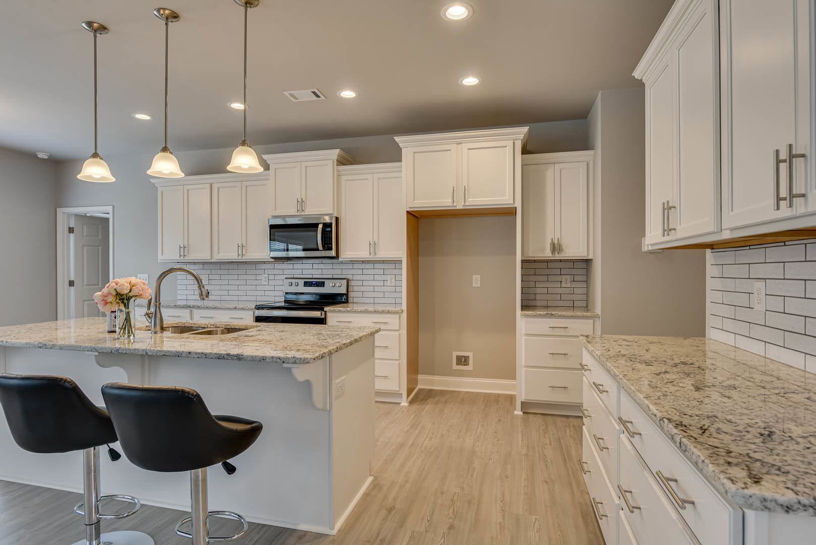 White kitchen cabinets with chrome bar stools at a marble countertop, pink flowers in a glass vase, built-in microwave, drawers, and exposed brick backsplash