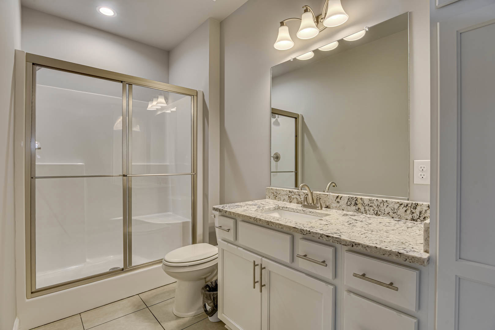 Modern bathroom featuring a glass-enclosed shower, white ceramic sink set in a stone countertop, wood cabinetry with drawers, chrome faucet, wall-mounted light fixture, and tile