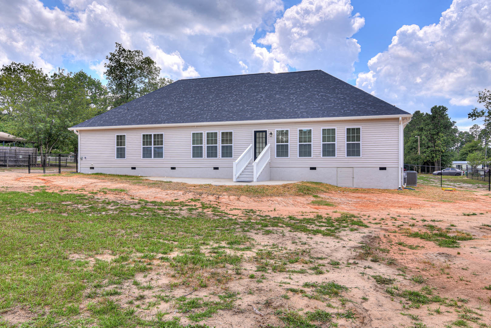 Two-story home with white siding, large windows, and white staircase, surrounded by a grassy yard and dirt field; heat pump unit visible near the foundation, roof set against a