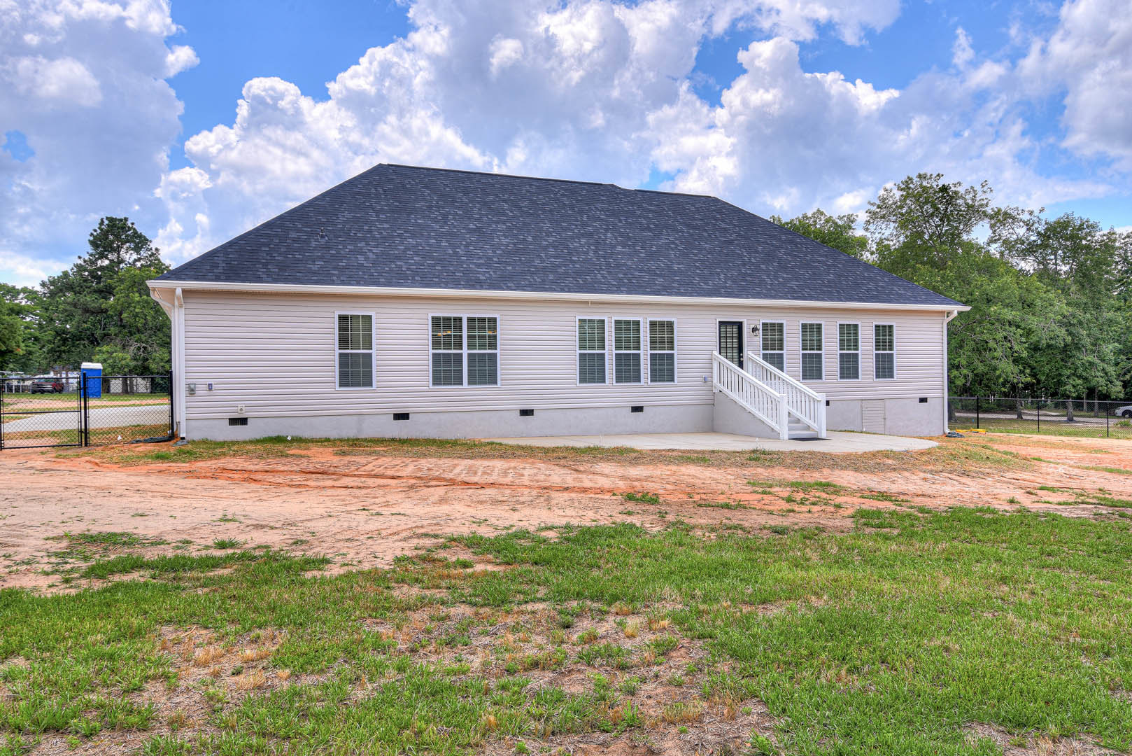 Blue-roofed house with white railings, surrounded by a dirt yard and patches of grass, under a partly cloudy sky