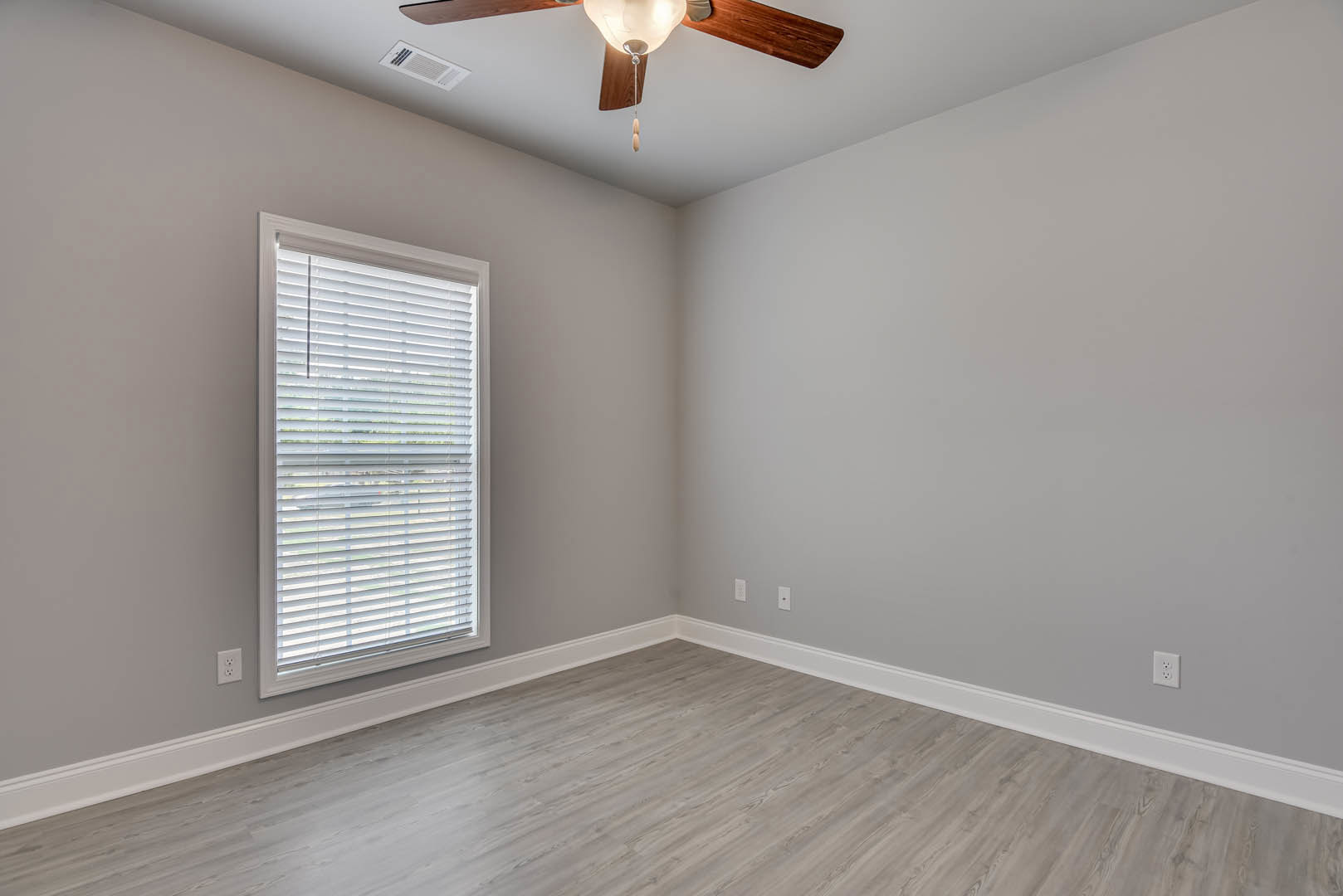 Wood flooring, white walls, ceiling fan with light fixture, large window with white trim, rectangular vent on wall, simple molding along ceiling and baseboards