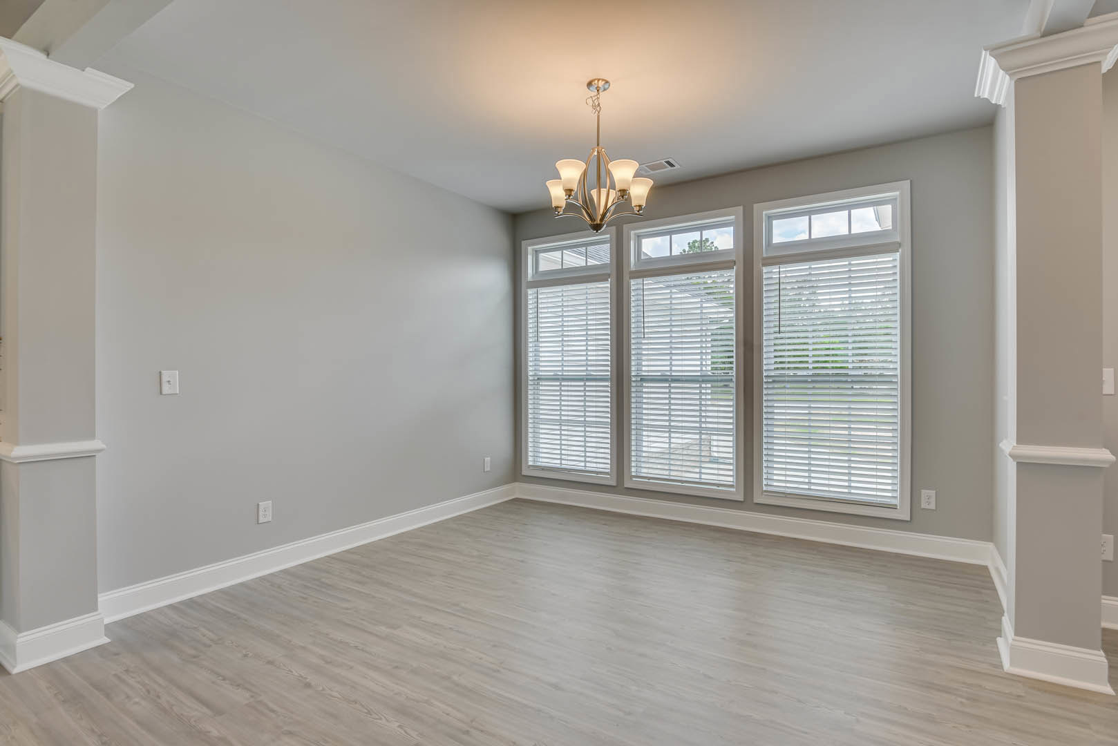 Chandelier hanging from ceiling above wood flooring, large windows with white blinds, white walls with decorative molding