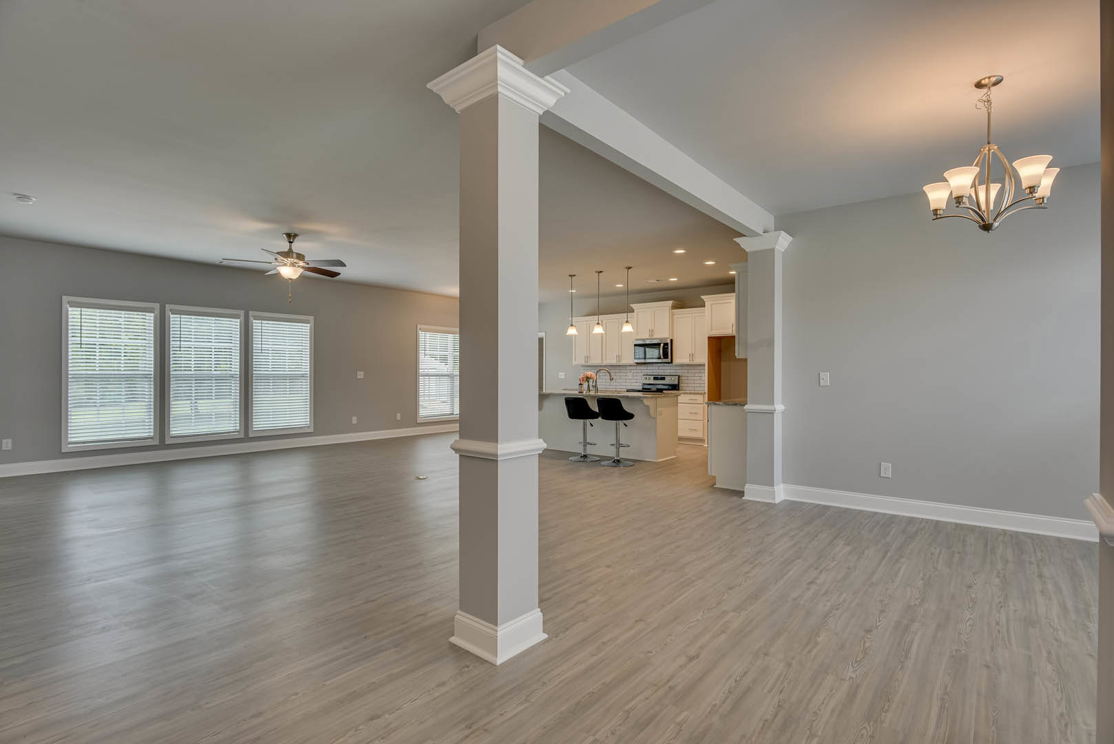 Spacious open floor plan featuring kitchen and dining area, wood laminate flooring, row of windows with blinds, two black chairs at kitchen counter, five-light fixture above
