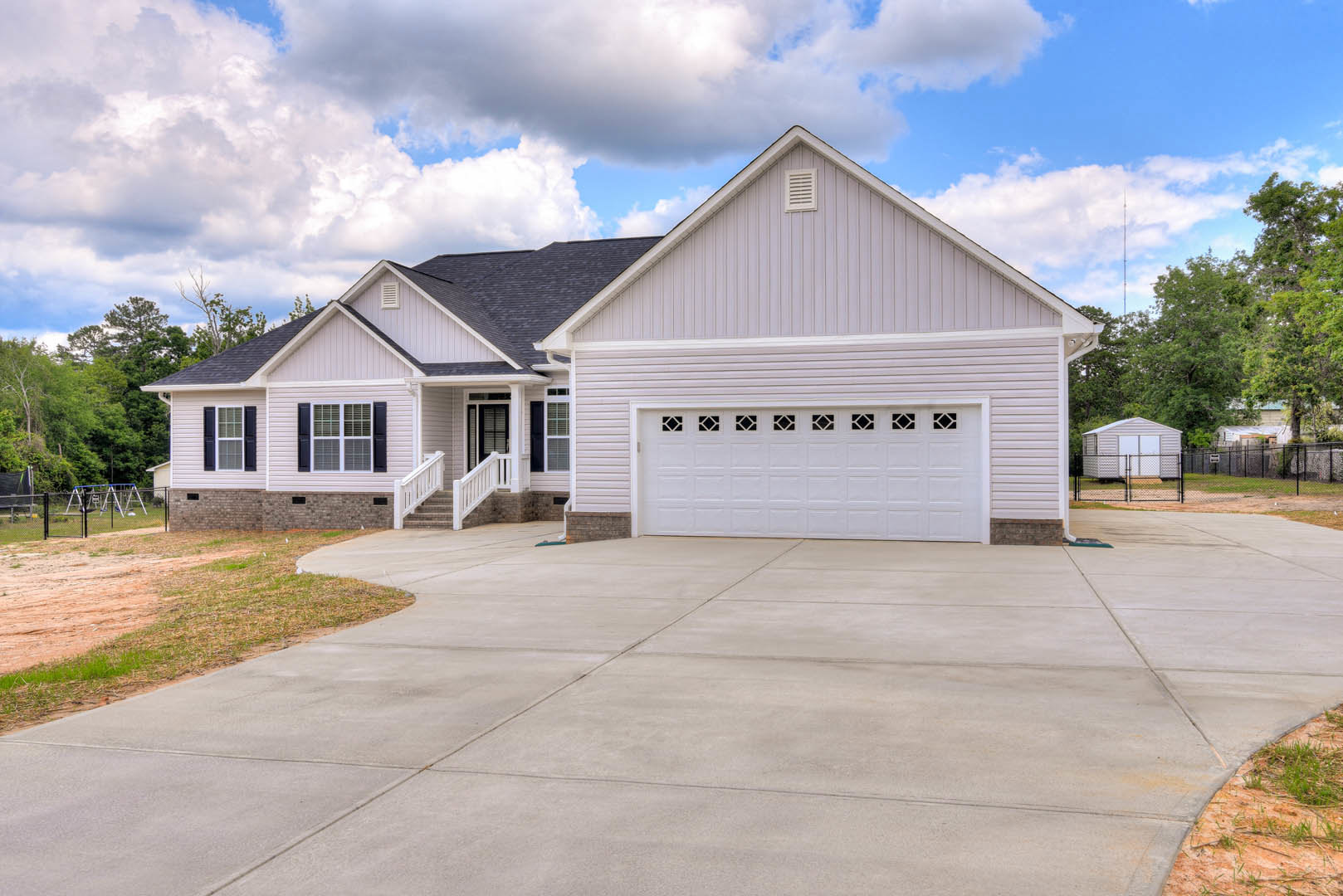 Two-story house with gray siding, white trim, concrete driveway leading to a single garage door, front steps, window with dark shutters, and roof vent, surrounded by trees under a