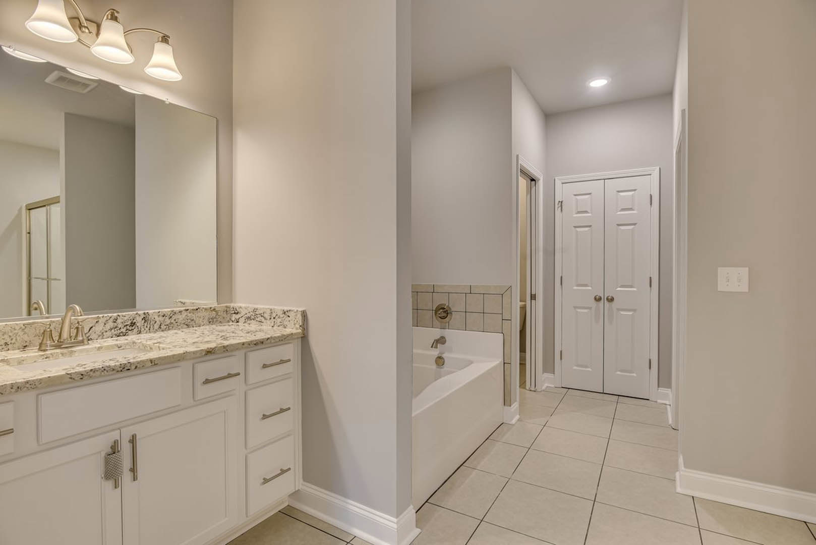 Bathroom with freestanding white bathtub and silver faucet, large wall mirror, white double doors with gold handles, ceiling light fixture, neutral walls and flooring