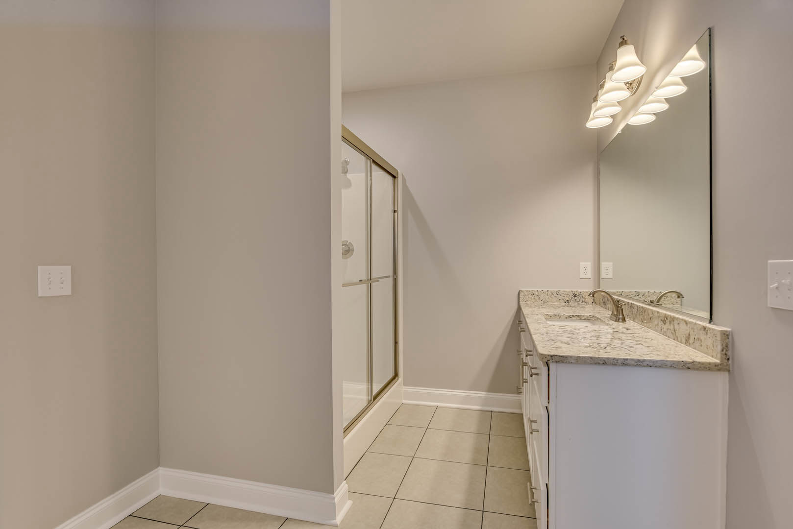 Bathroom with marble countertops, glass-enclosed shower, row of ceiling lights, white sink, and light switch on white plaster wall