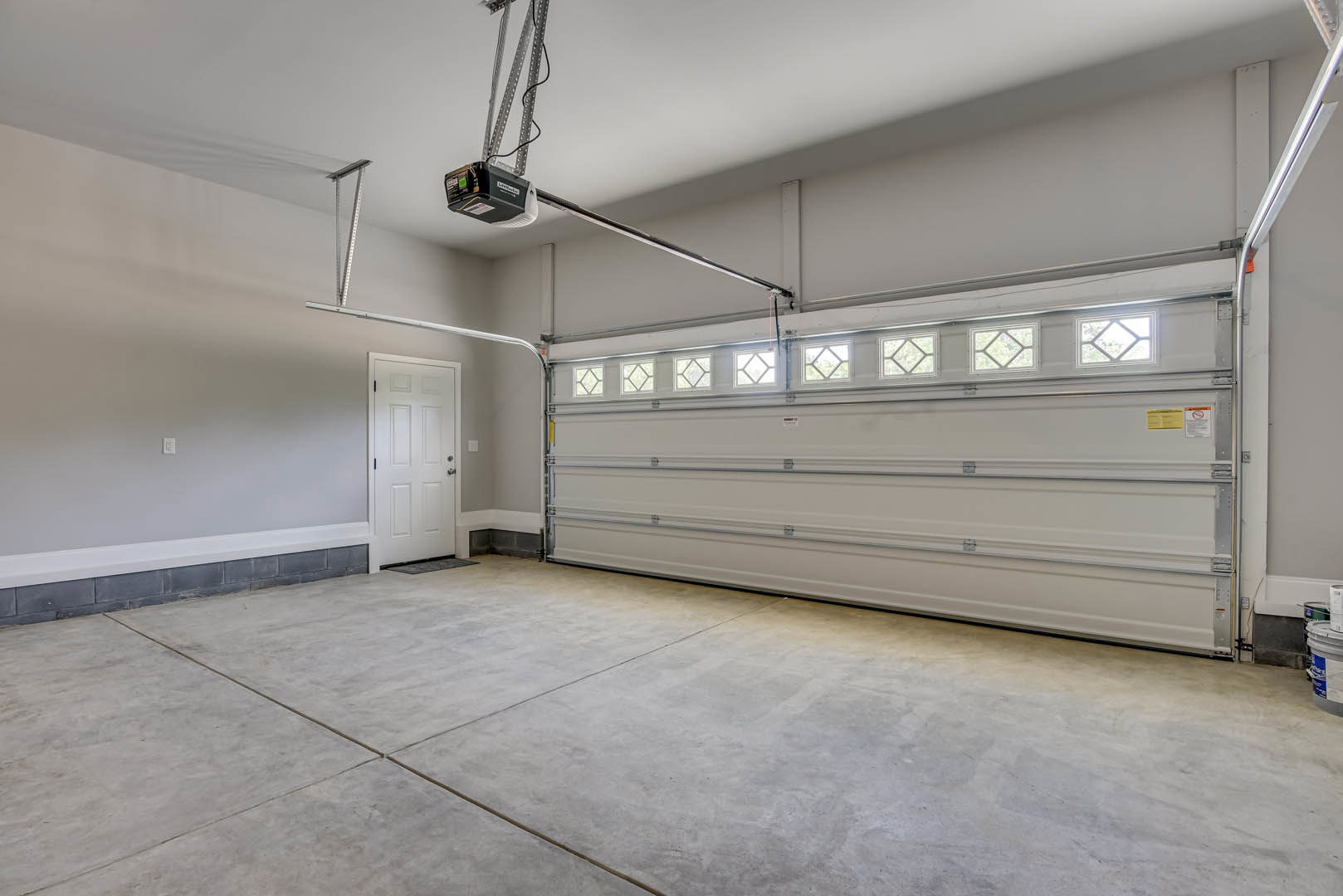 Spacious garage with concrete floor, large white door featuring silver handle, multiple windows, wall-mounted light fixture, and plaster walls.
