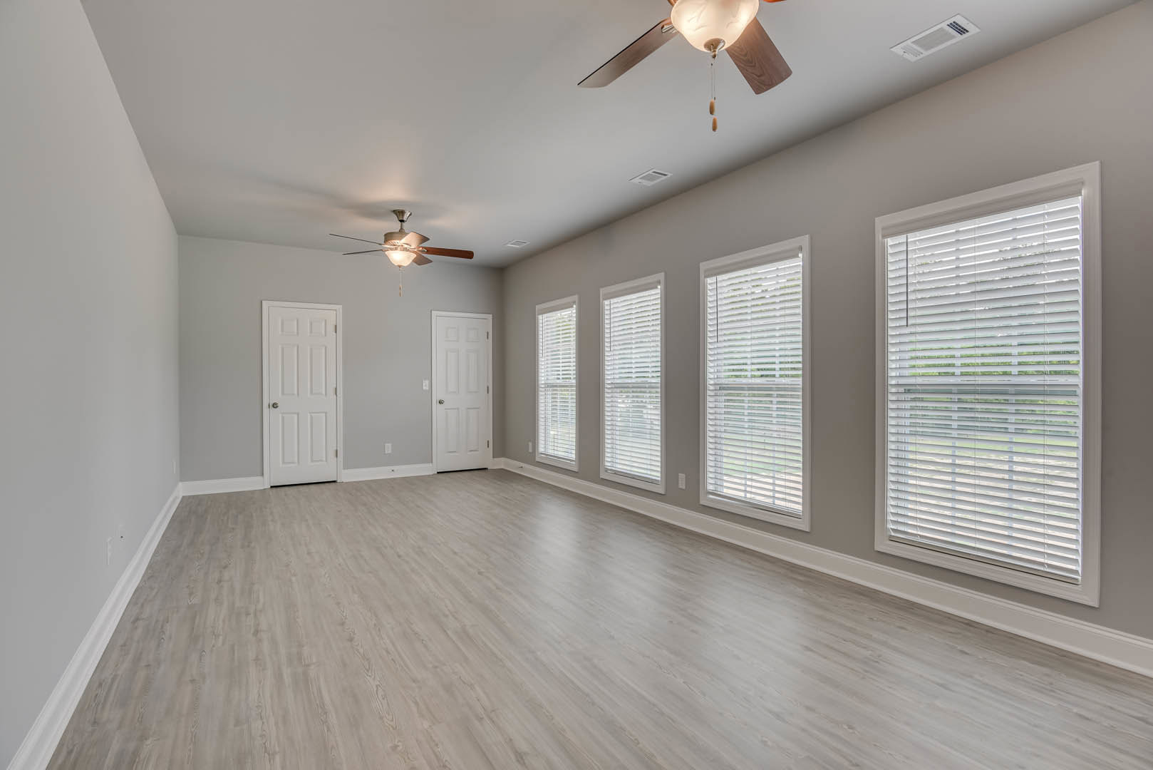 Neutral-toned room with laminate wood flooring, white plaster walls, ceiling fan, white door with silver knob, and windows fitted with blinds