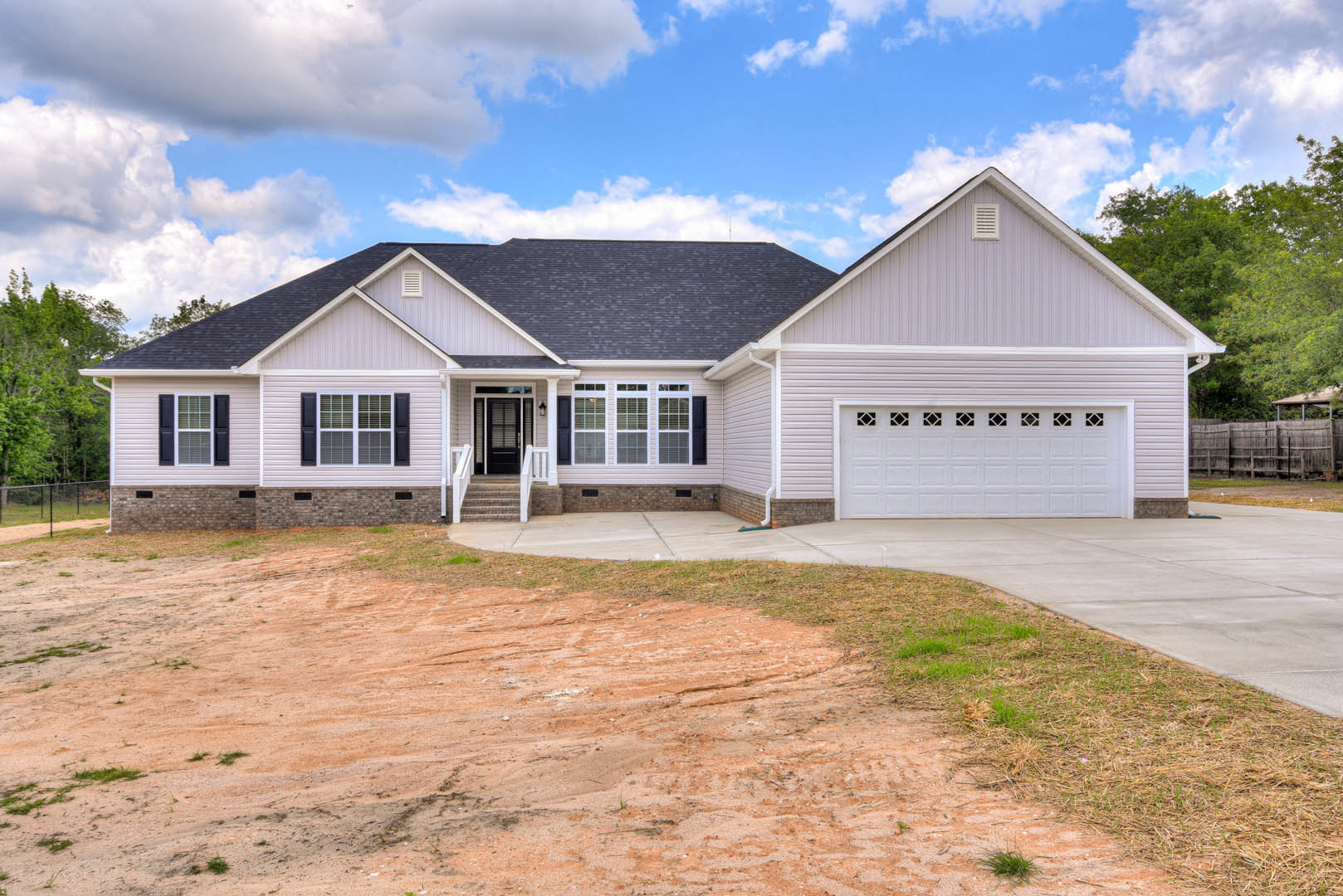 Two-story home with gray siding, white trim, covered front porch, concrete driveway, and garage door; dirt patch and stairs leading to entry; white grid window visible.