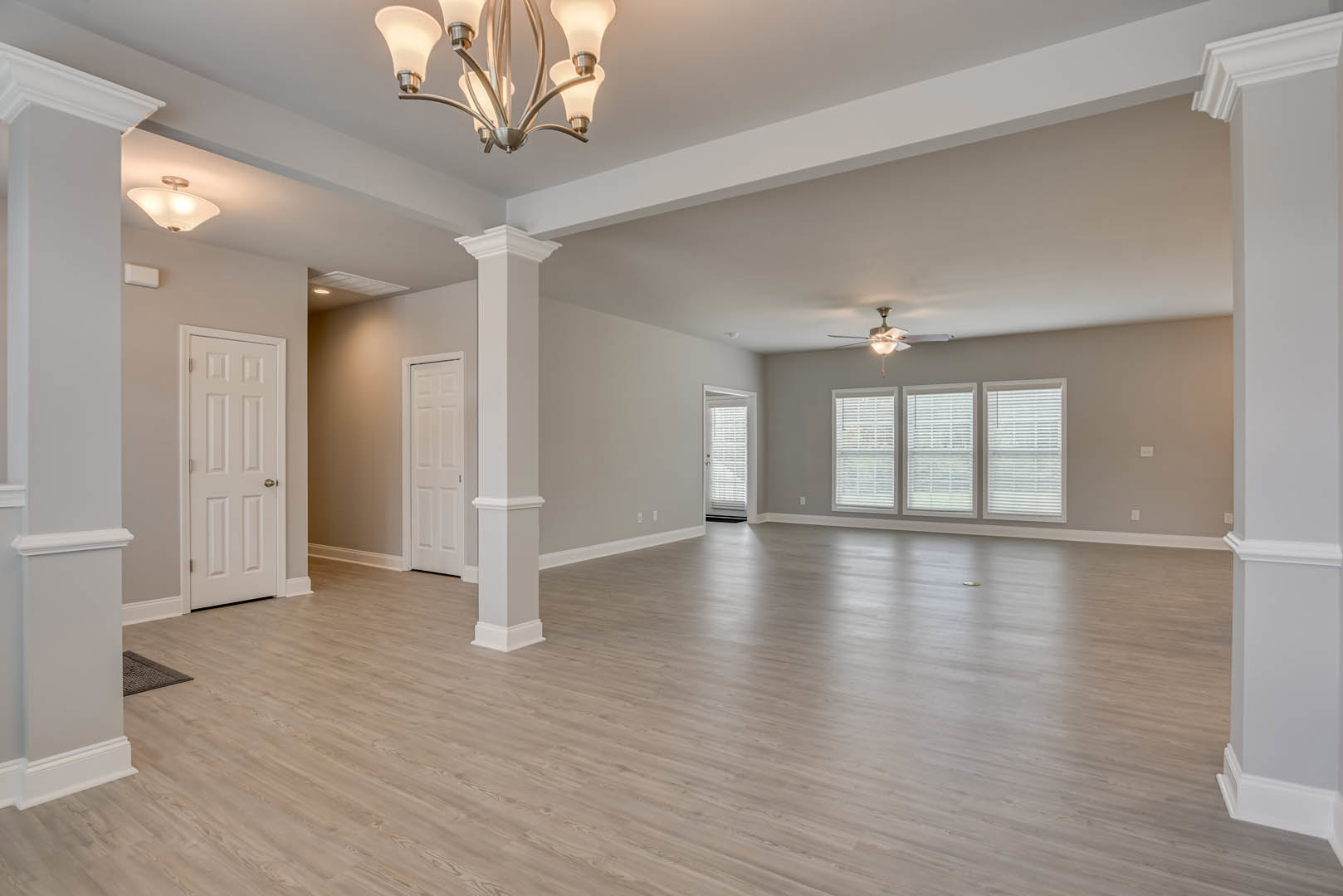 Spacious room with wood flooring, ceiling fan, ornate chandelier, row of windows with white blinds, white door featuring silver handle, light fixture mounted on plaster ceiling