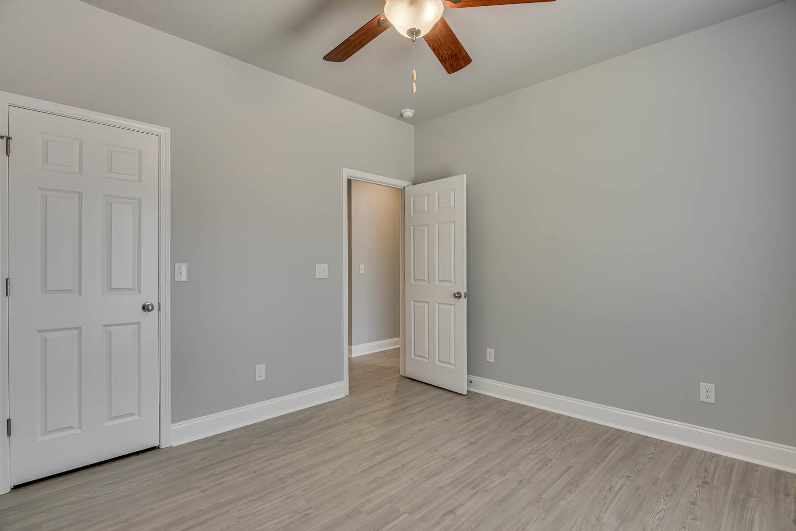 White paneled door with silver handle set in a room featuring light wood flooring, white plaster walls, ceiling fan with integrated light fixture, and nearby light switch.