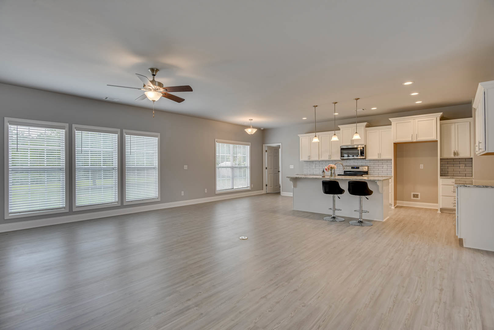 Spacious open floor plan featuring a kitchen with wood cabinetry, laminate flooring, dining area, ceiling fan with light, multiple windows with blinds, and neutral plaster walls