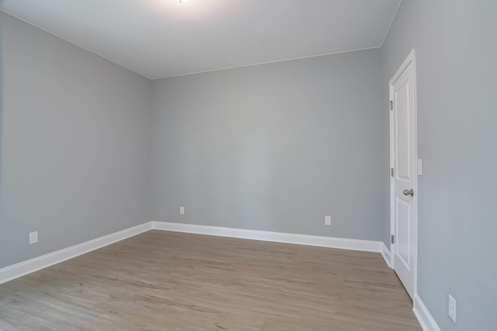 Wood flooring with white baseboard trim, white door featuring a silver handle, white walls, wall-mounted light fixture, and a rectangular object with a black border.