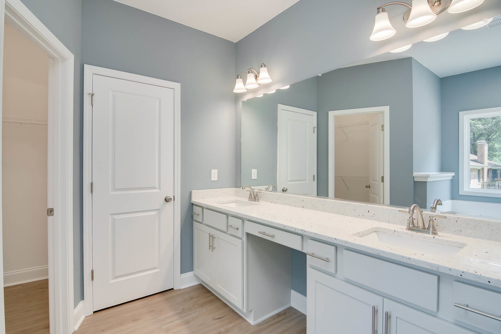 Bathroom with double vanity featuring white sinks, quartz countertop, large framed mirror, white cabinetry with silver hardware, light fixture with white shade, and white door with