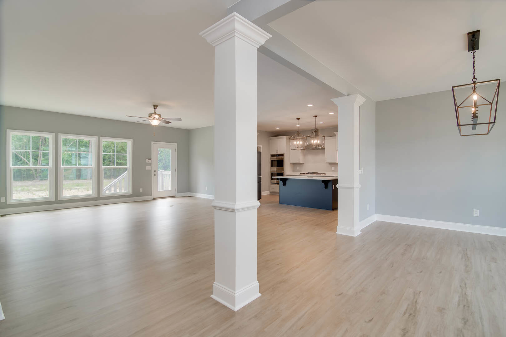 Open floor plan featuring a kitchen with wood flooring, white column, blue accent wall with a black fixture, ceiling light suspended by chain, and a row of windows providing