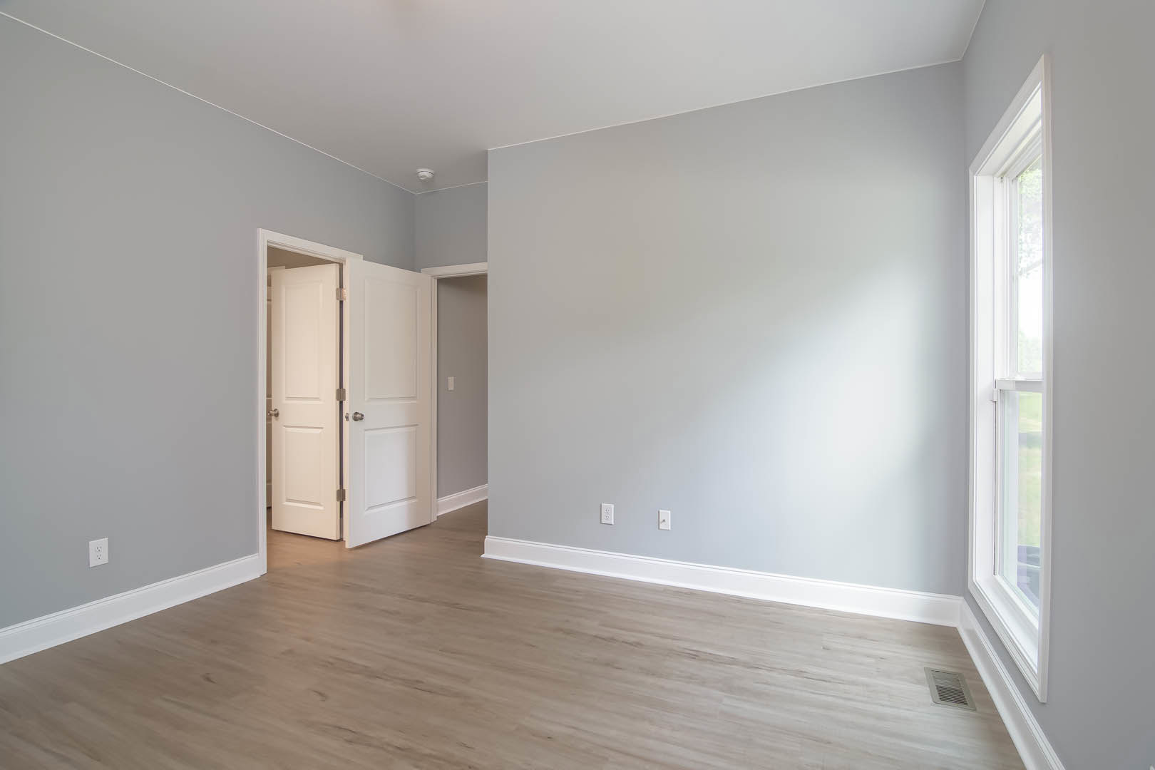 Hardwood floor with white baseboards, open white door with silver knobs, white walls, wall vent, and white-framed window in a residential room