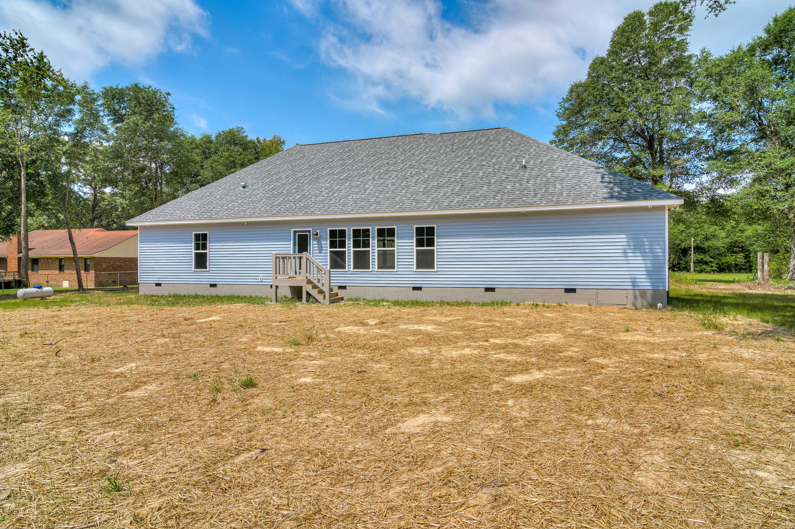 Two-story house with light siding, square-paned windows, wooden exterior staircase, straw-covered yard, mature trees in background, cloudy sky overhead