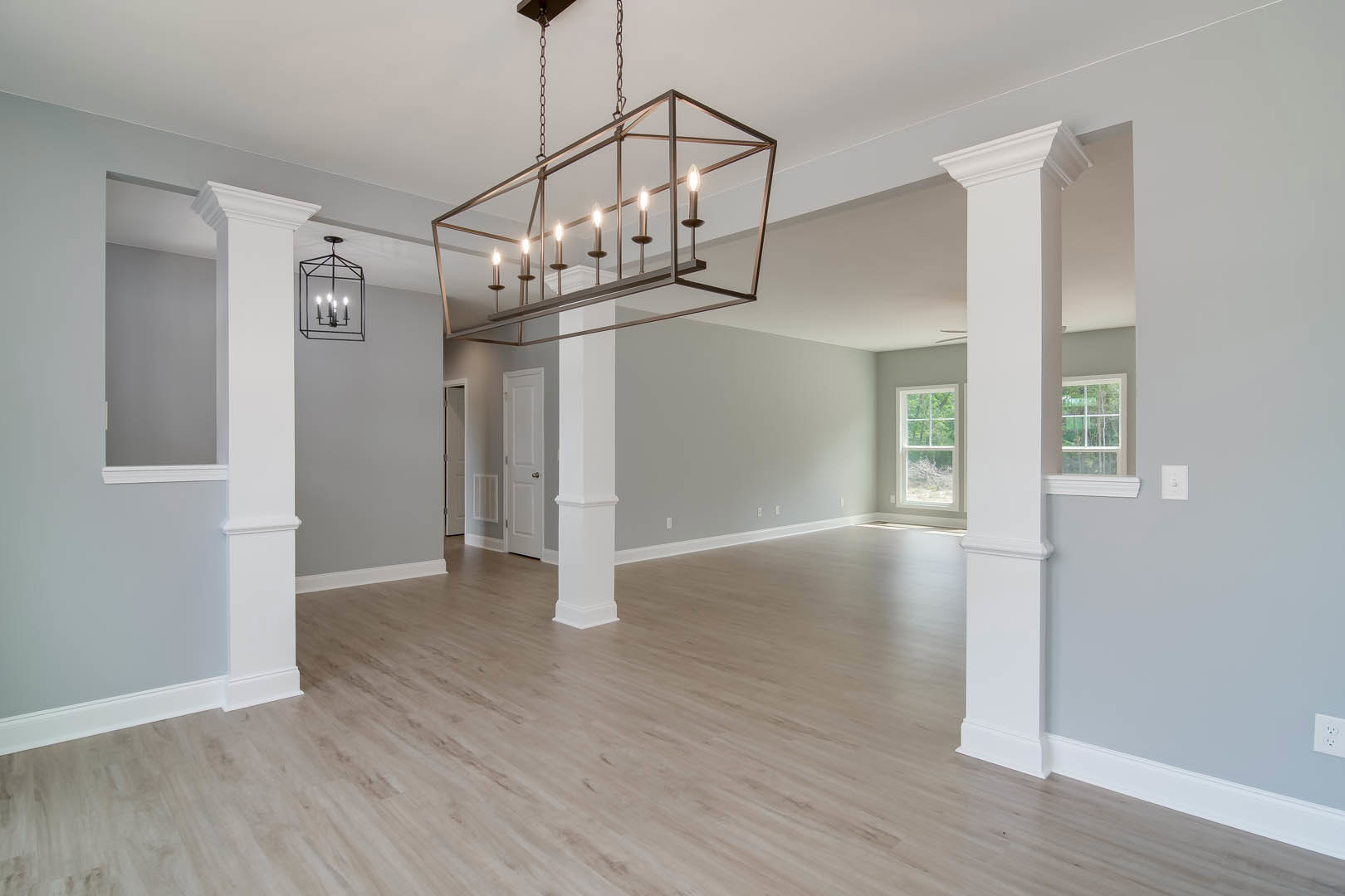 Open living space with white columns, rectangular chandelier, wood flooring, white walls, large window overlooking trees, and white door with silver knob