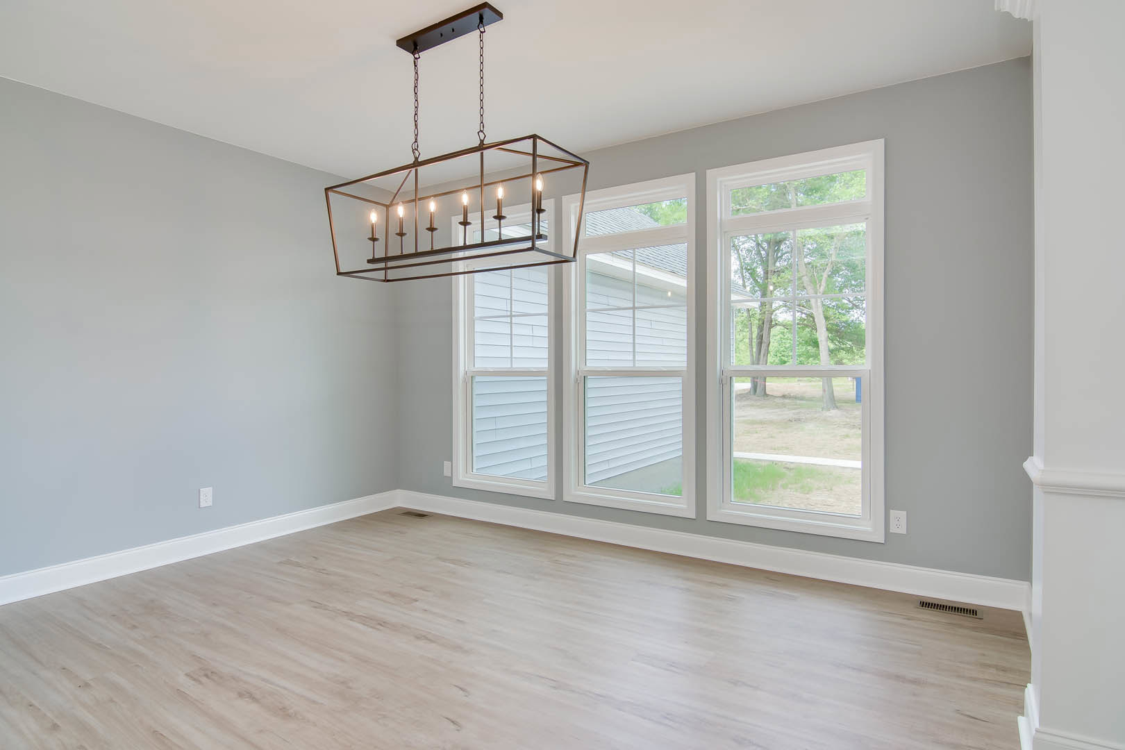 Hardwood floor room with large windows overlooking trees, ornate chandelier hanging from ceiling, white plaster walls, black chain supporting rectangular light fixture