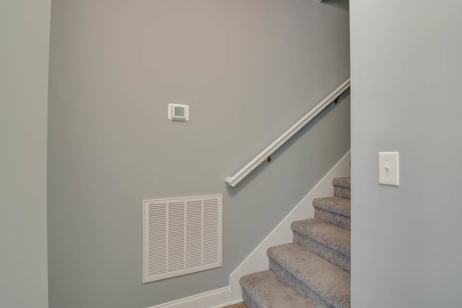 Wood staircase with white-painted handrail, plaster wall featuring a square white vent and light switch, hardwood floor at base.